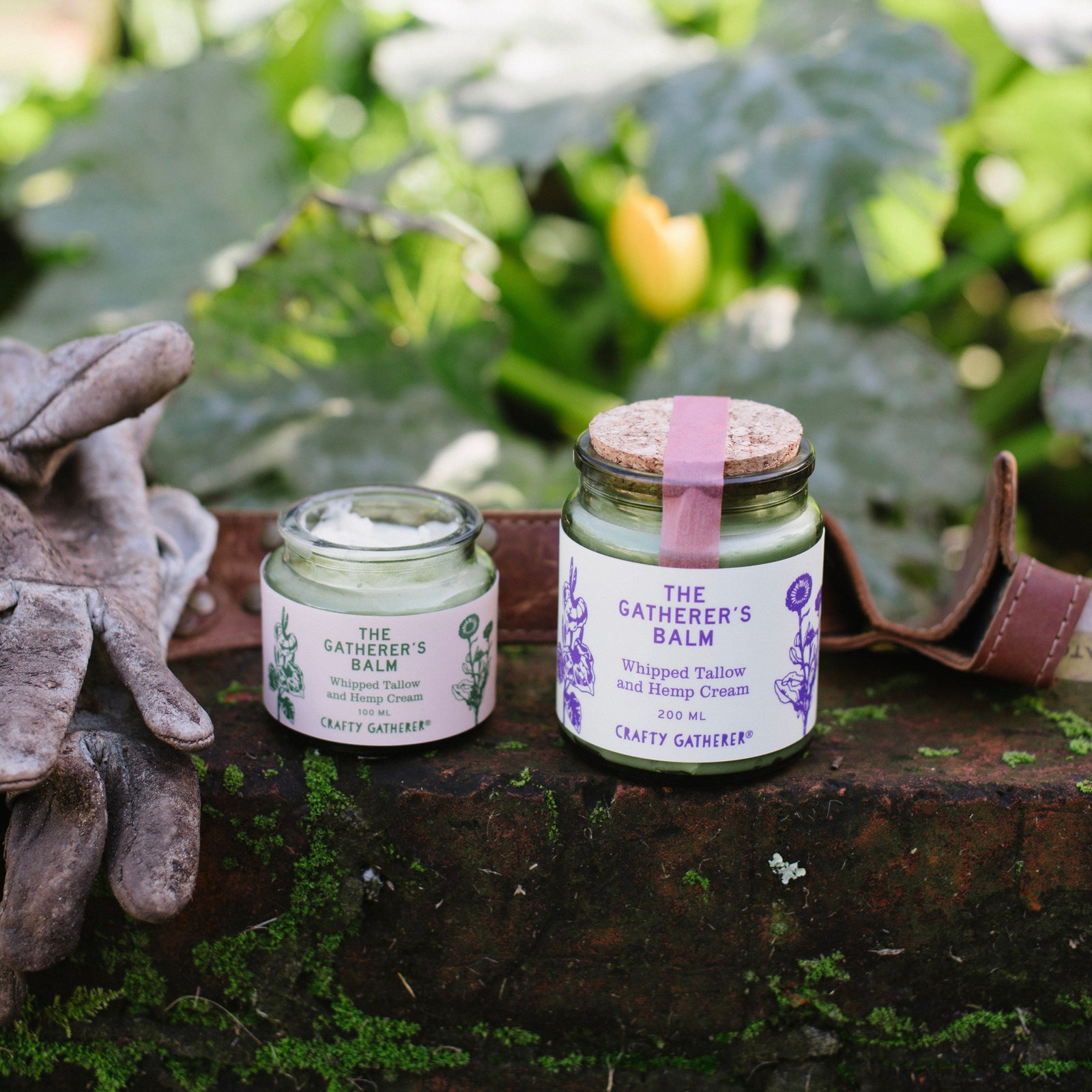 Two jars of 'The Gatherer's Balm' on a wooden surface with greenery in the background.