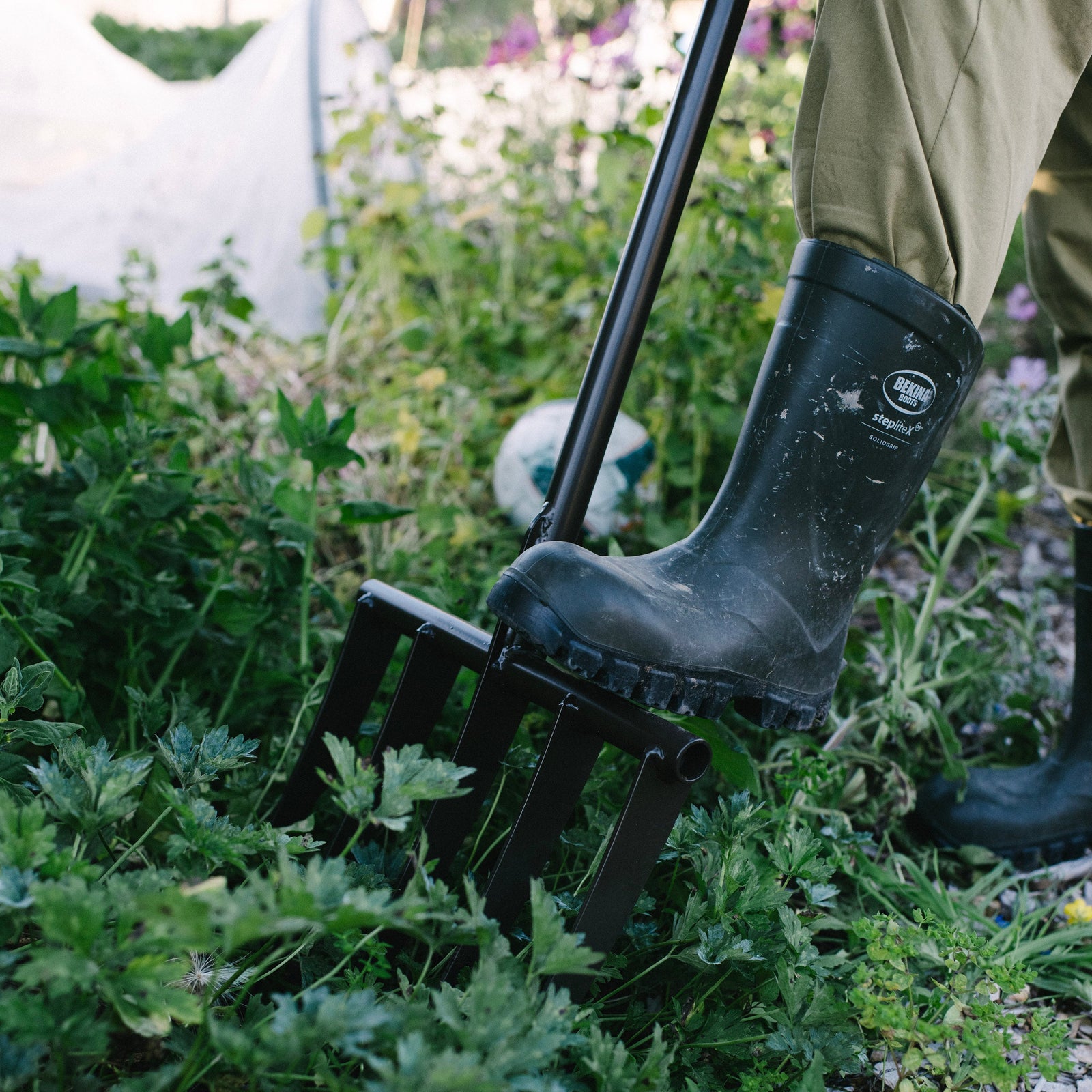 Person using a garden forksta in a garden setting with greenery and a boot visible.
