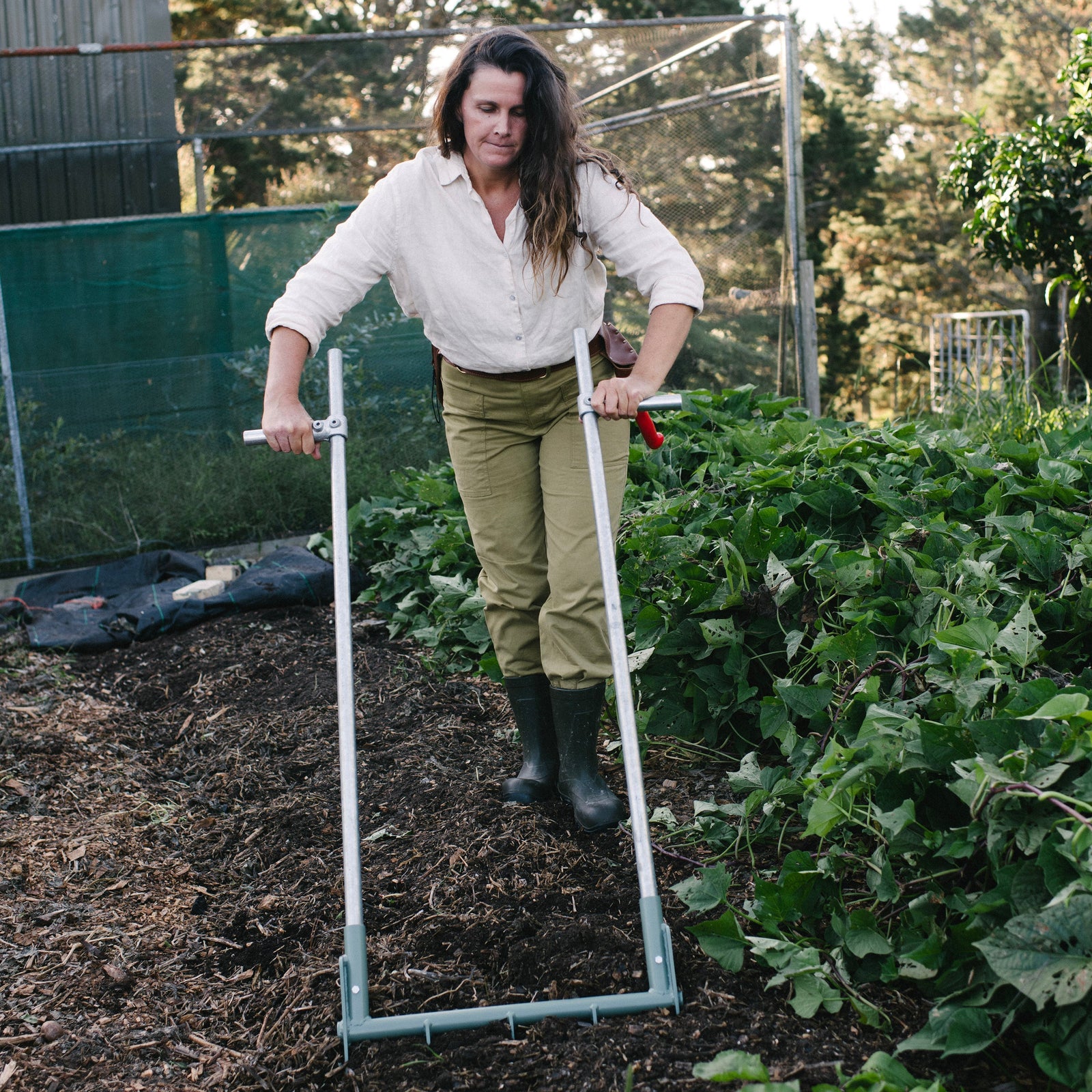 Person using a garden tool in an outdoor setting with greenery and trees. NZ made broadfork.