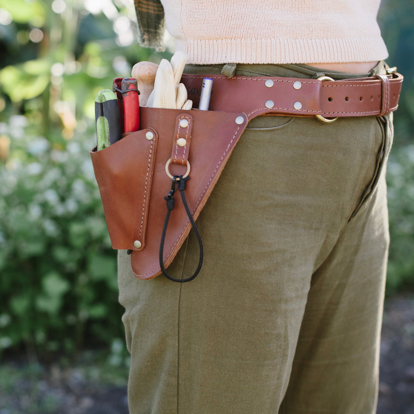 Person wearing a brown leather tool belt with various tools against a blurred natural background