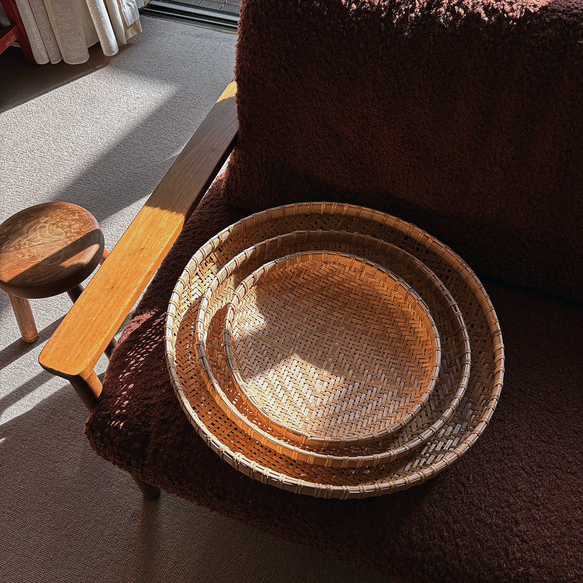 Three nested round woven rattan trays in natural tan tones, displayed on a dark brown textured armchair in a sunlit room.