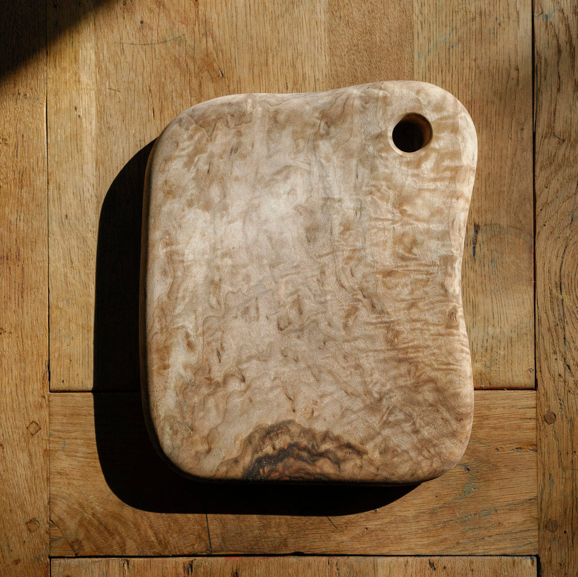 A rustic, hand-carved wooden cutting board with a natural wavy grain pattern and a small hole in the top right corner, displayed on a matching wooden plank surface.