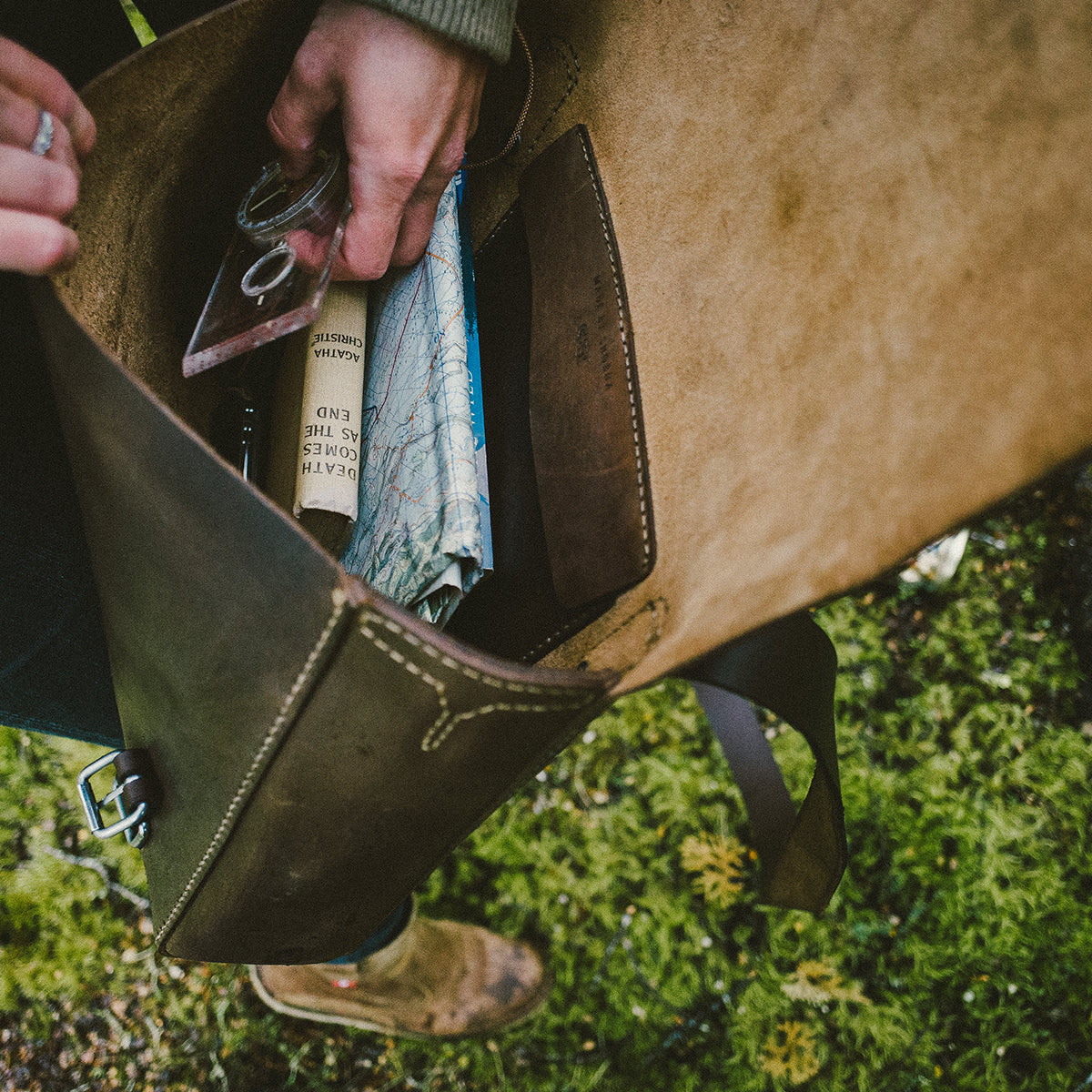 Brown leather satchel with items like a book and glasses on a green grass background