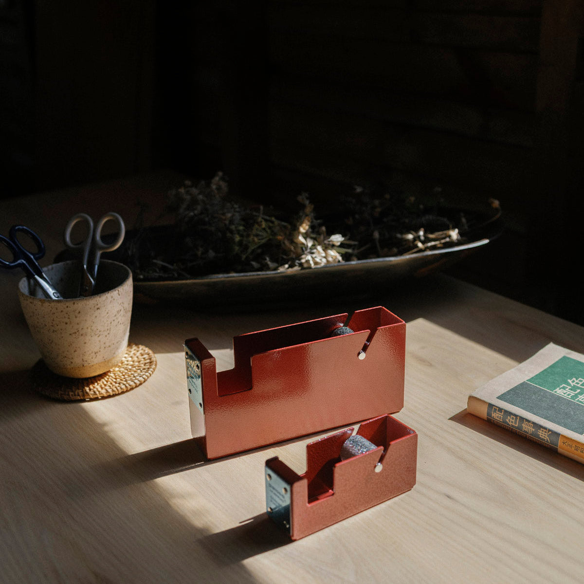 Red metal tape dispensers on a wooden surface with a cup and books in the background