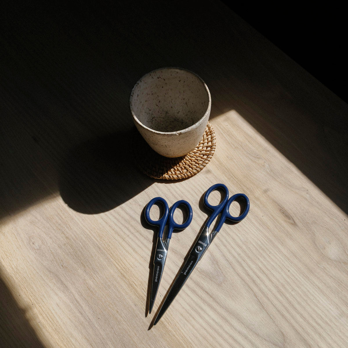 Two pairs of blue-handled scissors on a wooden surface with a ceramic cup in the background.