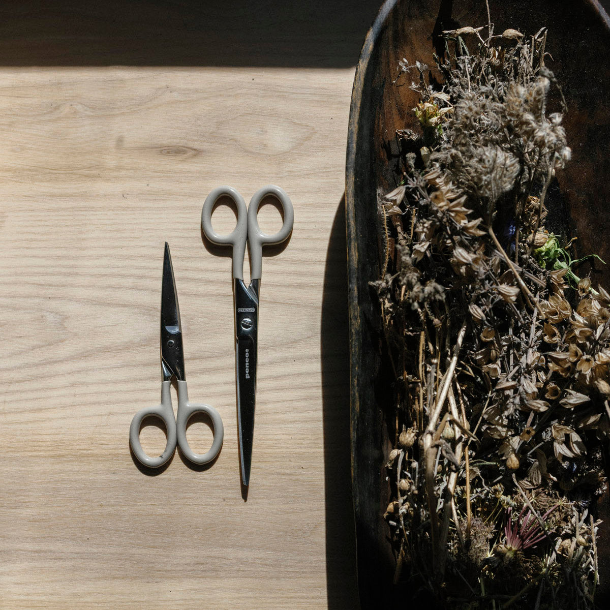 Two pairs of scissors on a wooden surface with a bowl of dried herbs.