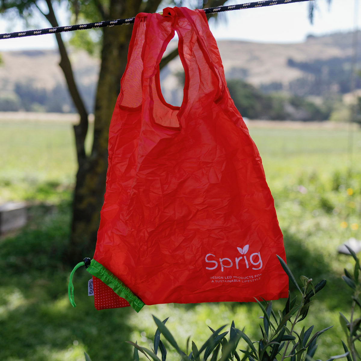 Red reusable shopping bag with 'Sprig' branding hanging on a tree branch outdoors.