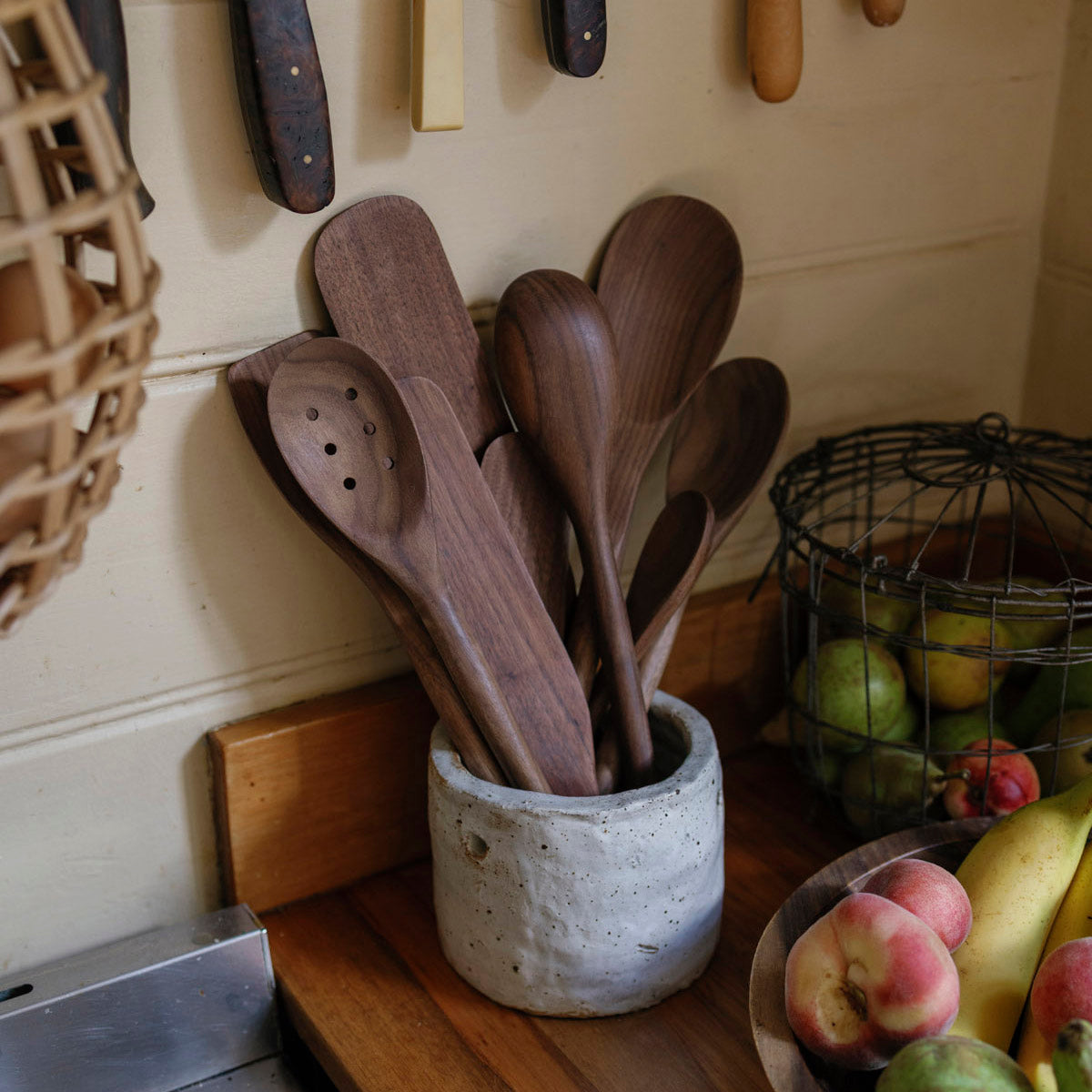 Set of walnut wooden spoons in a concrete holder on a kitchen counter with fruits and utensils in the background.