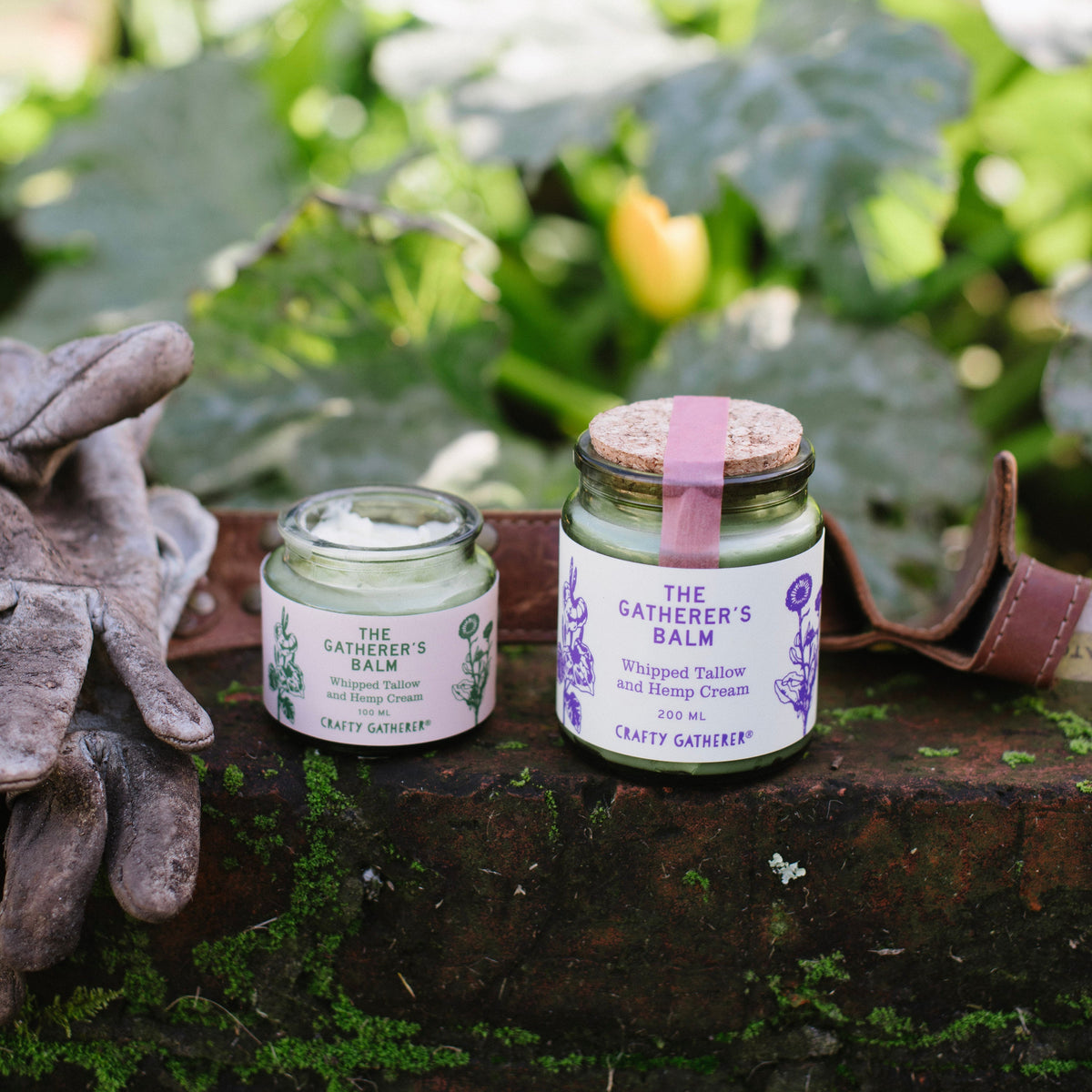 Two jars of 'The Gatherer's Balm' on a wooden surface with greenery in the background.