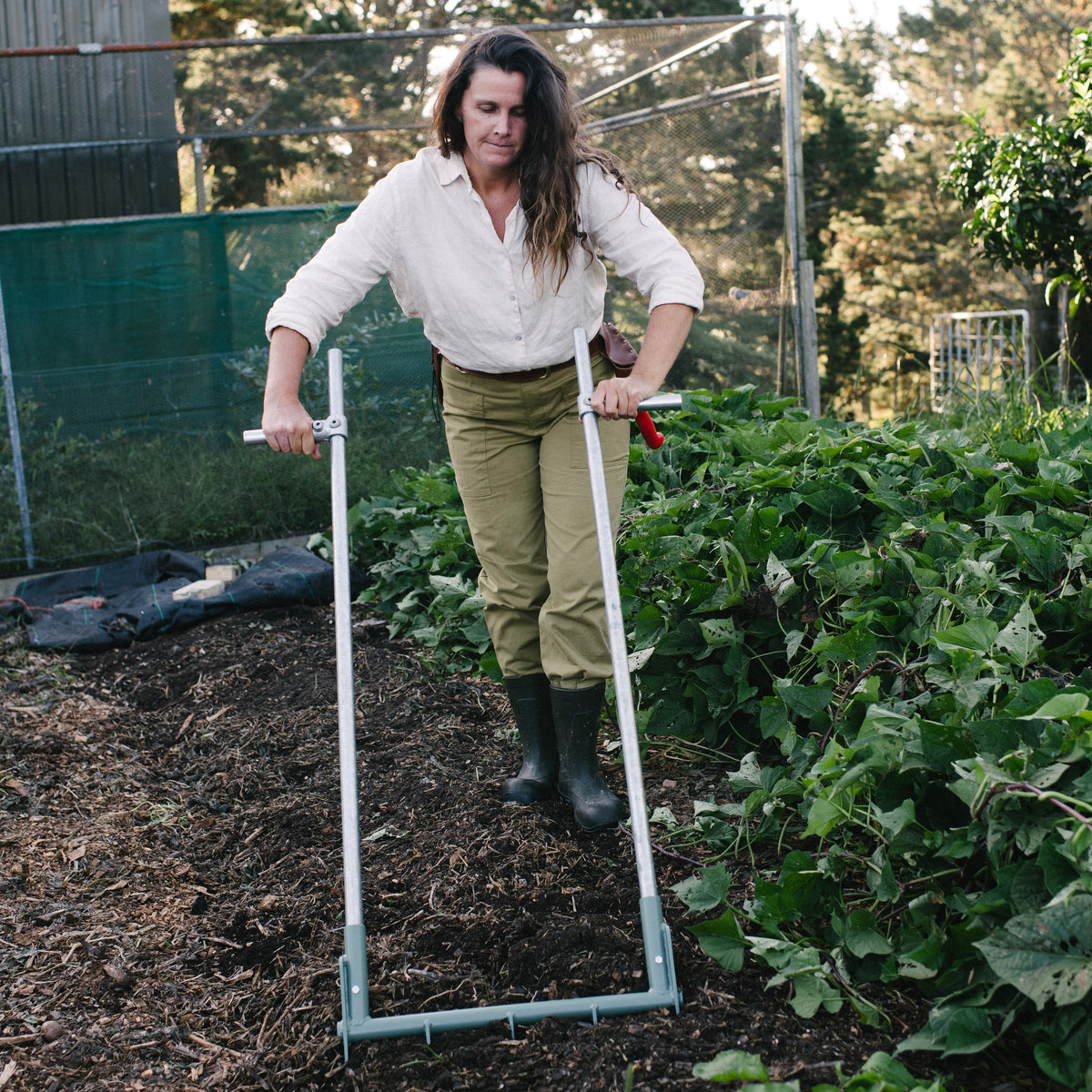 Person using a garden tool in an outdoor setting with greenery and trees. NZ made broadfork.