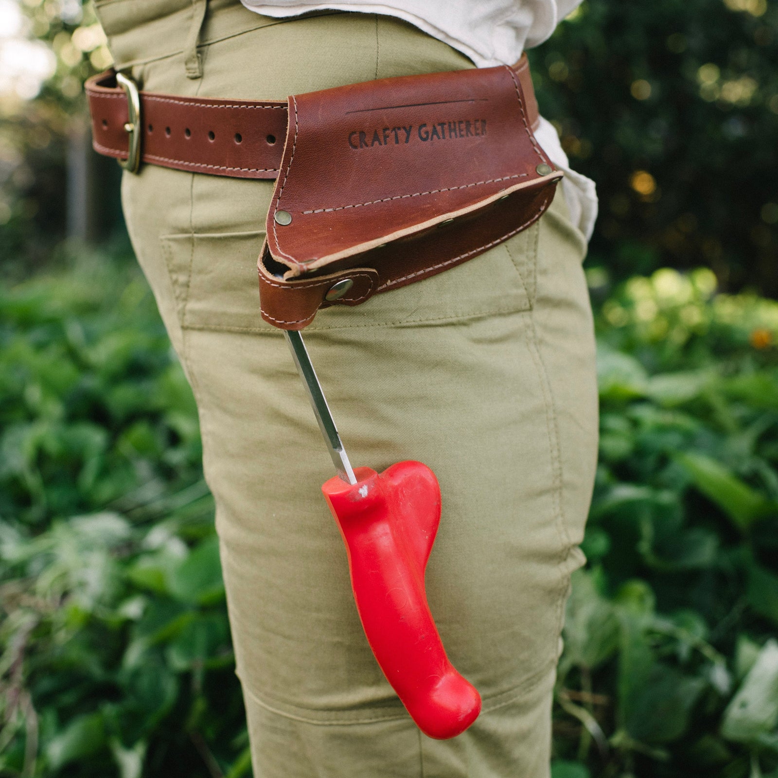 Person wearing a leather tool belt with a red tool, standing in a garden.