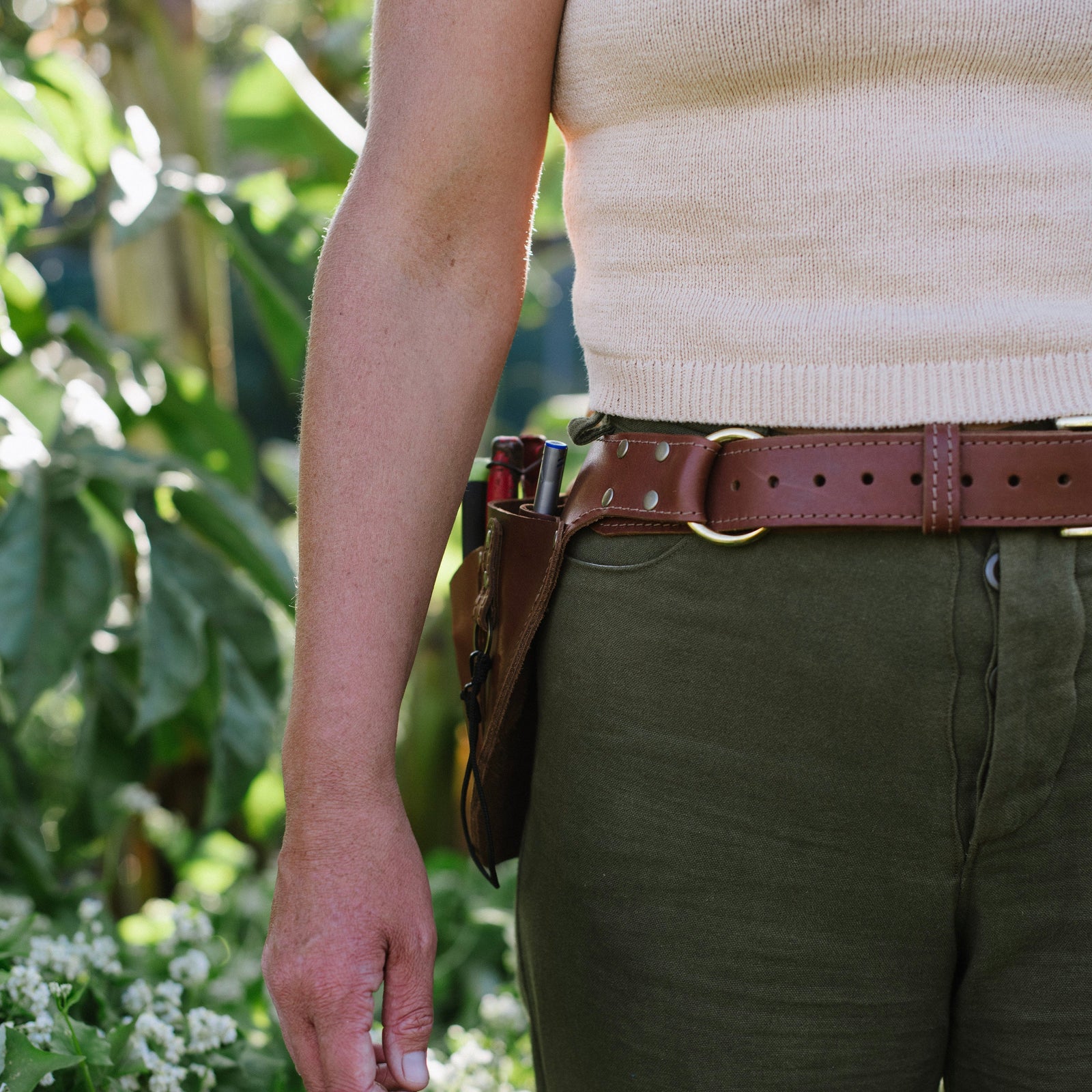 Person wearing a beige sleeveless top and green pants with a brown belt, standing in a garden.