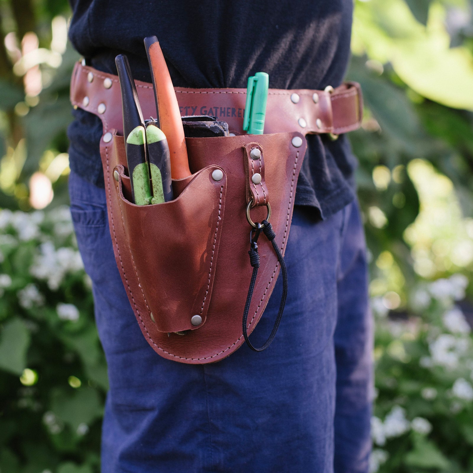 Person wearing a leather tool belt with gardening tools against a blurred green background