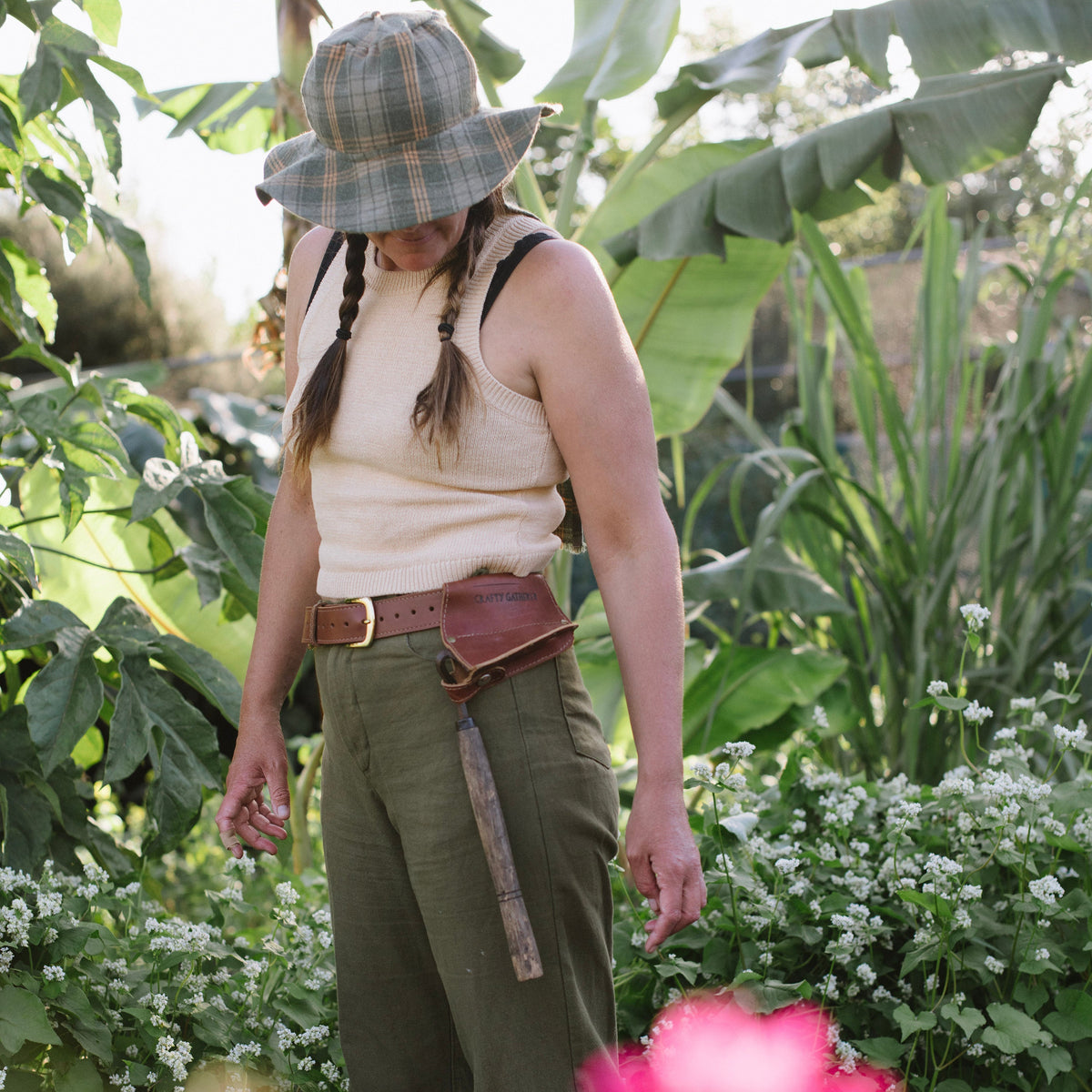 Person in a garden wearing a plaid hat, beige top, and green pants.