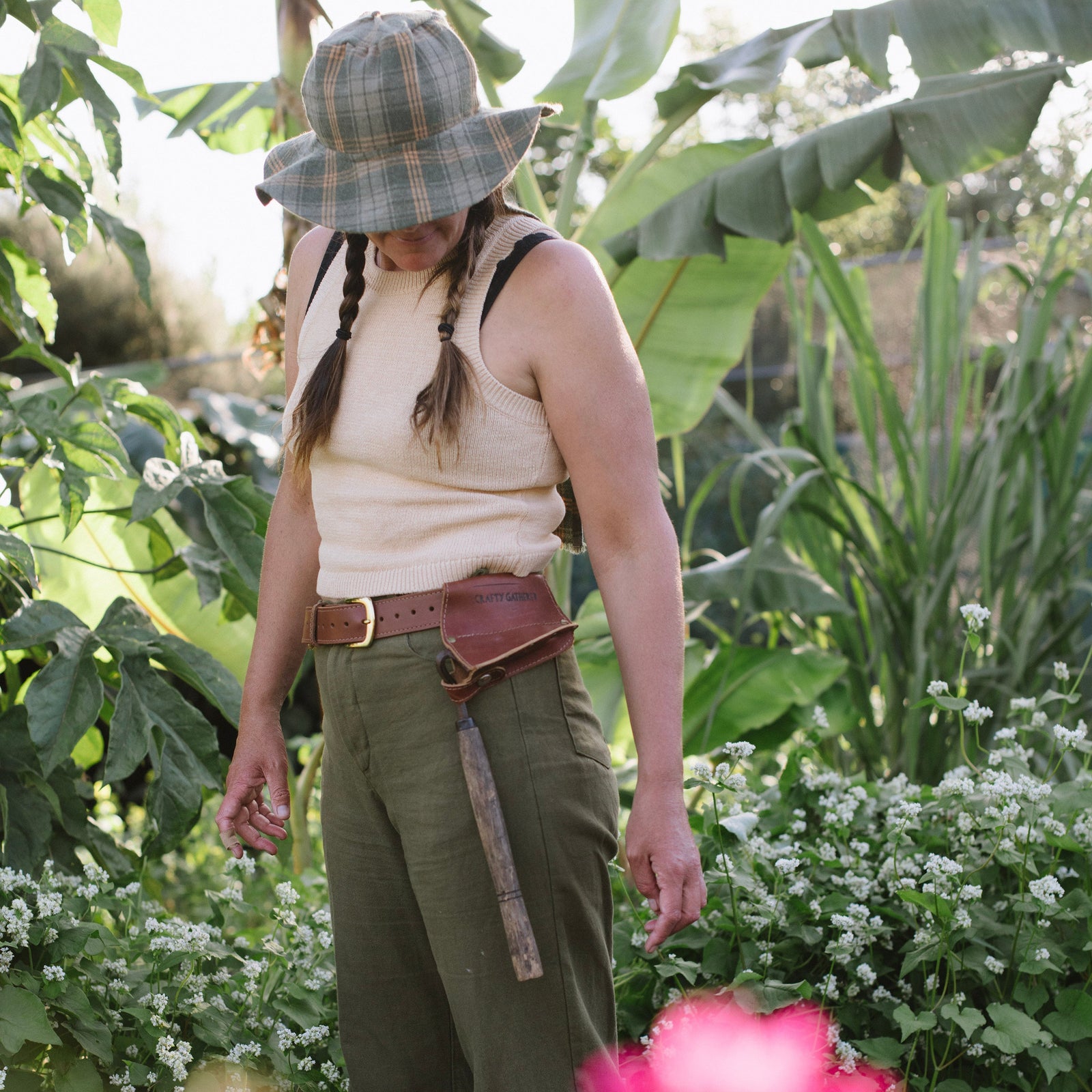 Person in a garden wearing a plaid hat, beige top, and green pants.