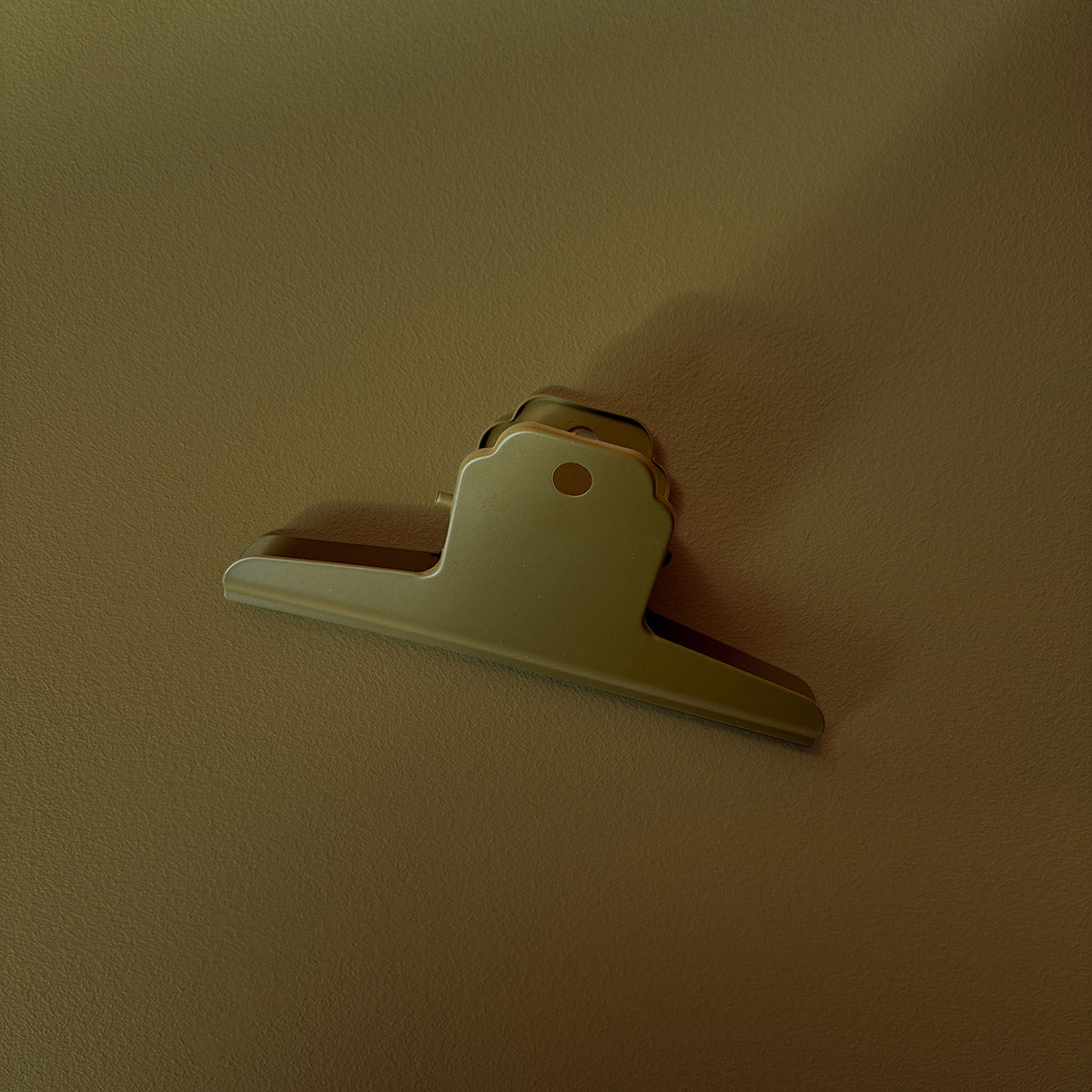 A green wall-mounted binder clip or hook with a small circular hole, photographed against a plain textured wall.