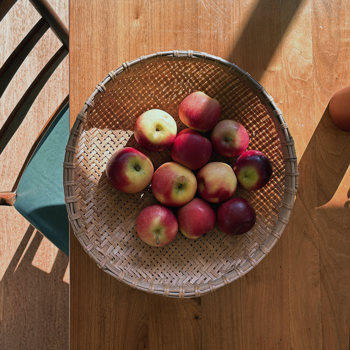 A round woven basket filled with red and yellow apples, placed on a wooden table in warm sunlight.