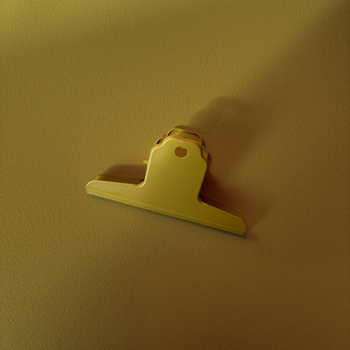 A small Chartreuse metal bulldog clip or fastener with a metal screw base, shown against a textured beige wall under warm lighting.