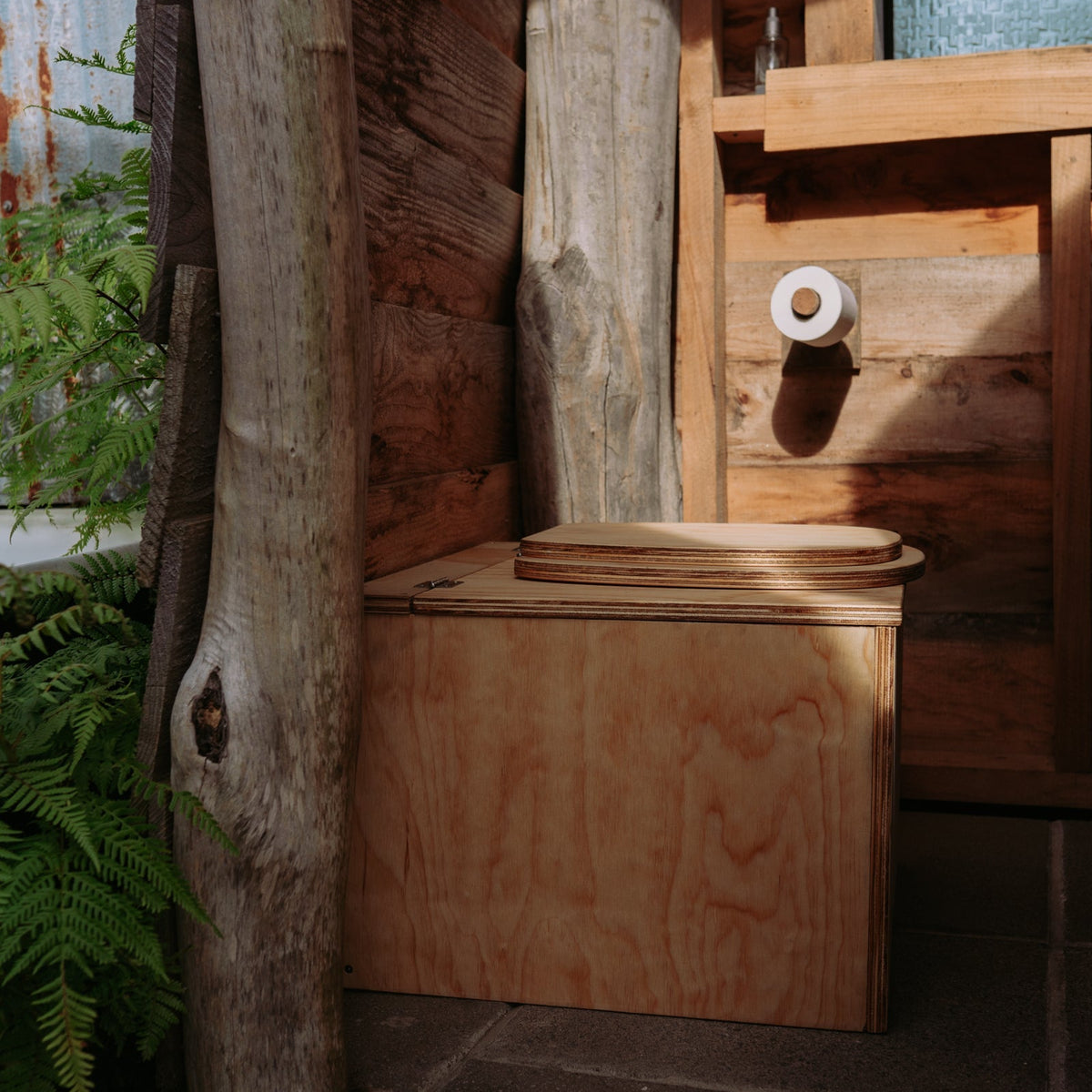 Wooden composting toilet in an outdoor setting with plants and a wooden structure.
