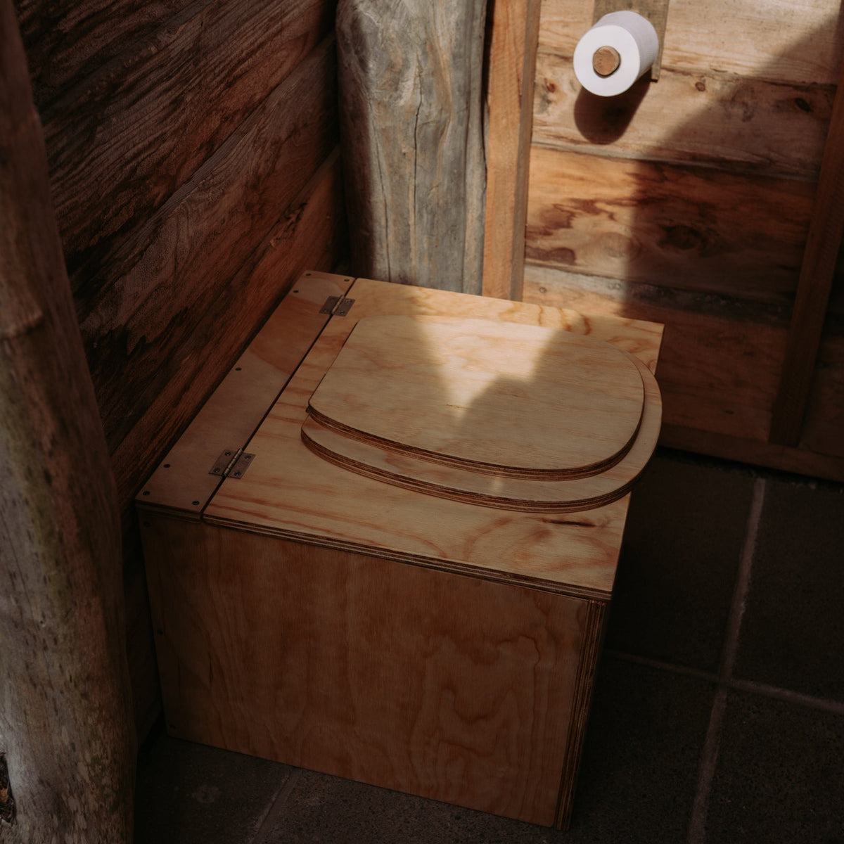 Wooden composting toilet in a rustic bathroom setting with a roll of toilet paper.