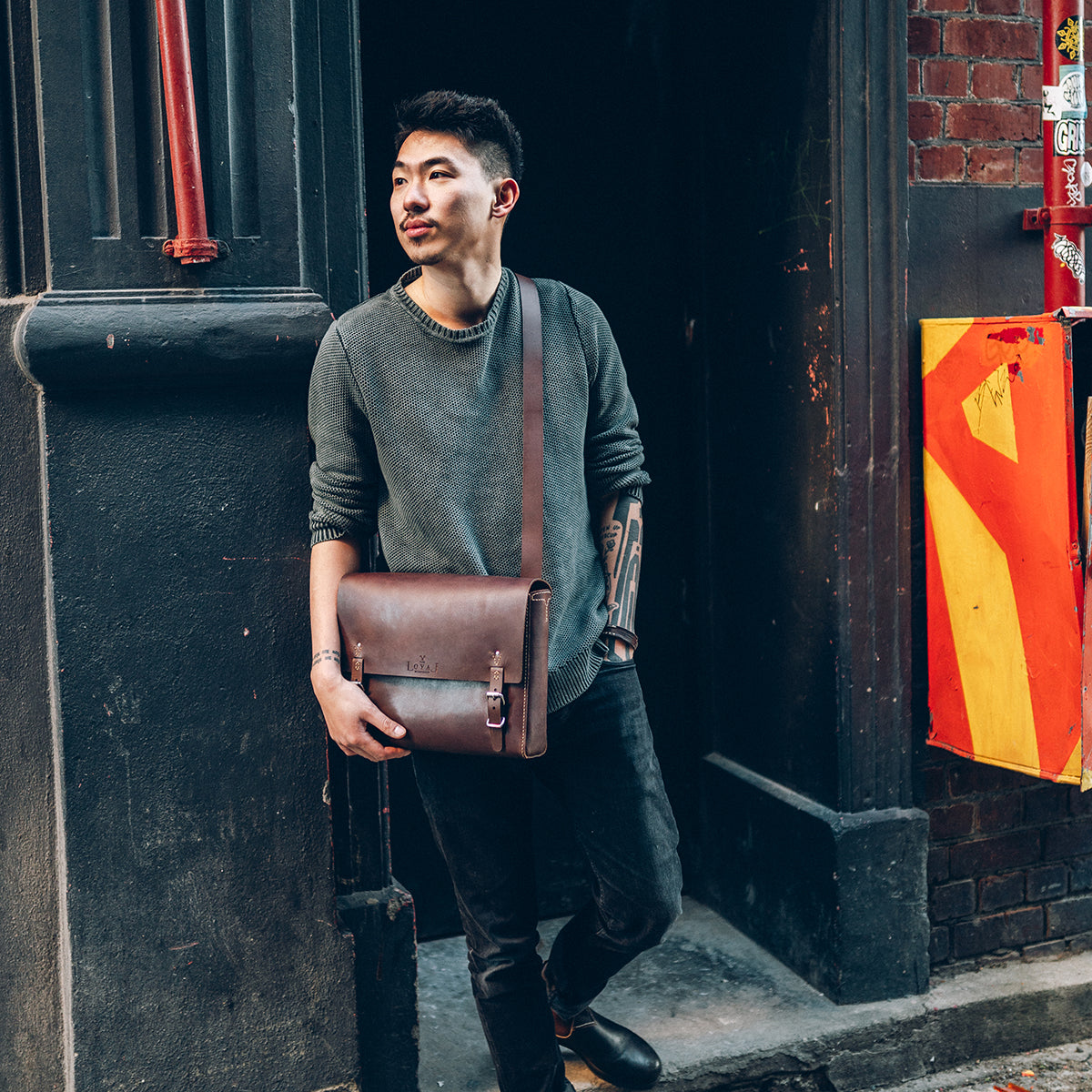 Man holding a brown leather satchel against a dark door with a red and yellow flag in the background.