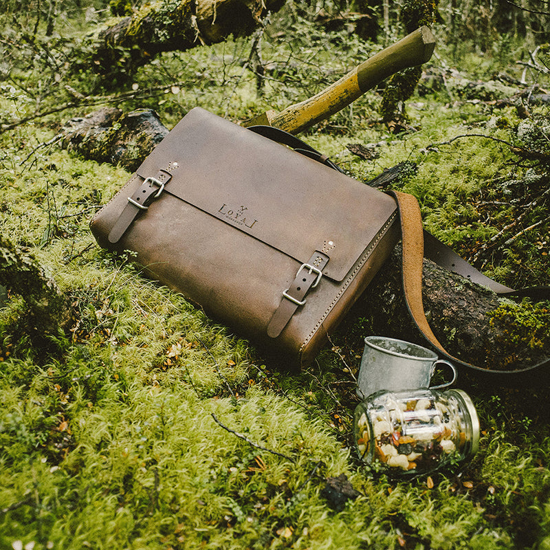 Brown leather satchel with a brand logo on a mossy forest floor.