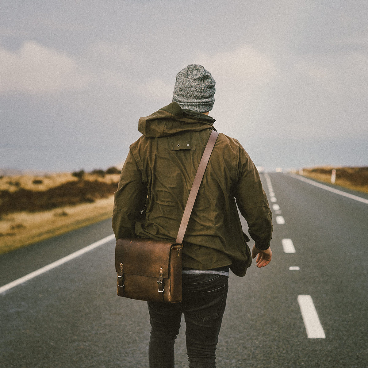 Person walking alone on a road wearing a green jacket and carrying a brown satchel.