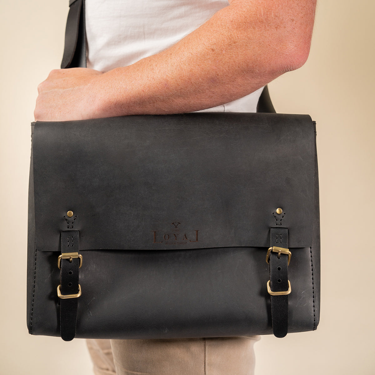 Person holding a black leather satchel bag with gold buckles on a beige background