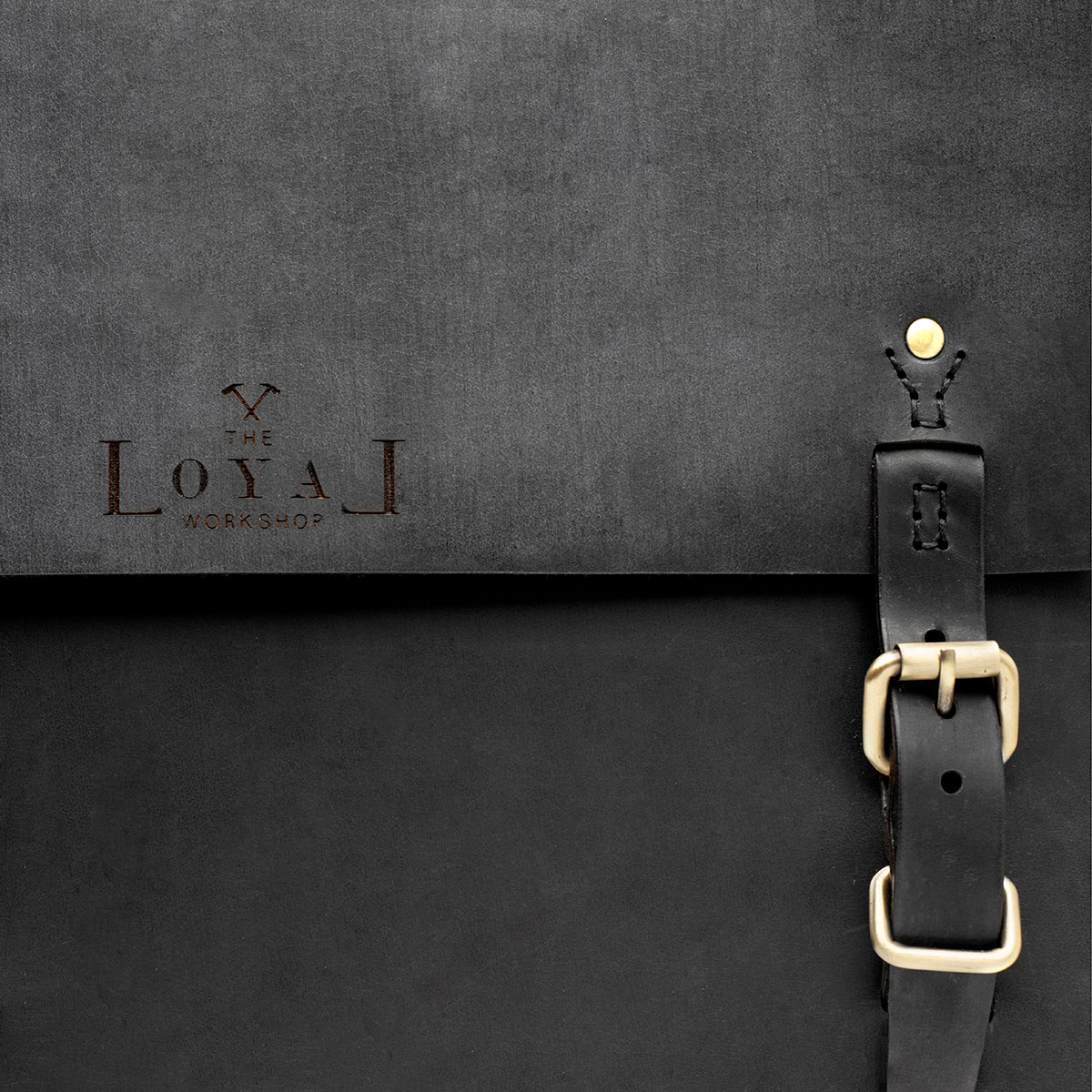Black leather bag with gold buckle on a dark gray background, featuring &#39;The Loyal Workshop&#39; logo.