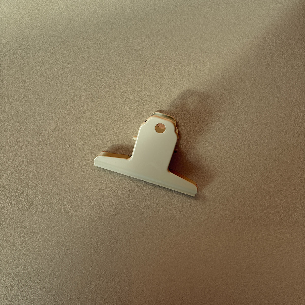 A small white metal bulldog clip with a gold-toned metal spring and backing, photographed against a textured beige wall with soft shadows.