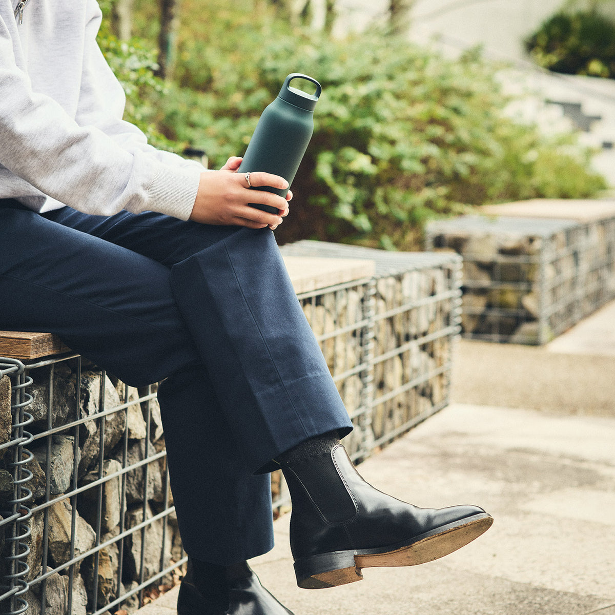 Person sitting outdoors holding a green water bottle, wearing dark boots and blue pants.