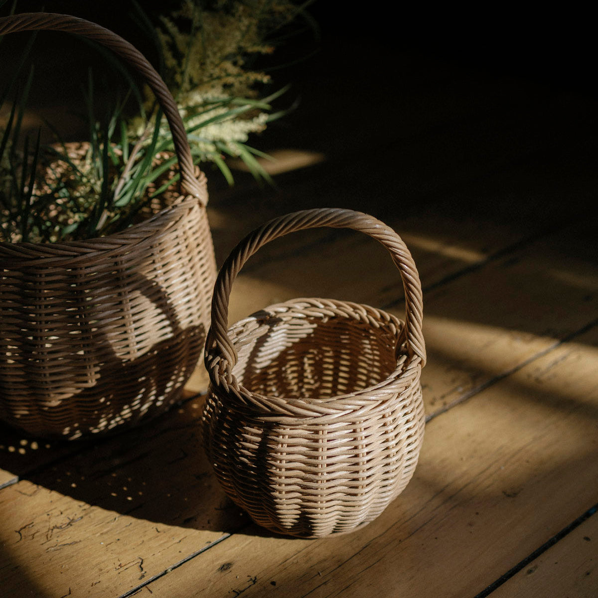 Two woven wicker baskets with handles, one small and one larger, placed on a wooden floor in dramatic warm sunlight.