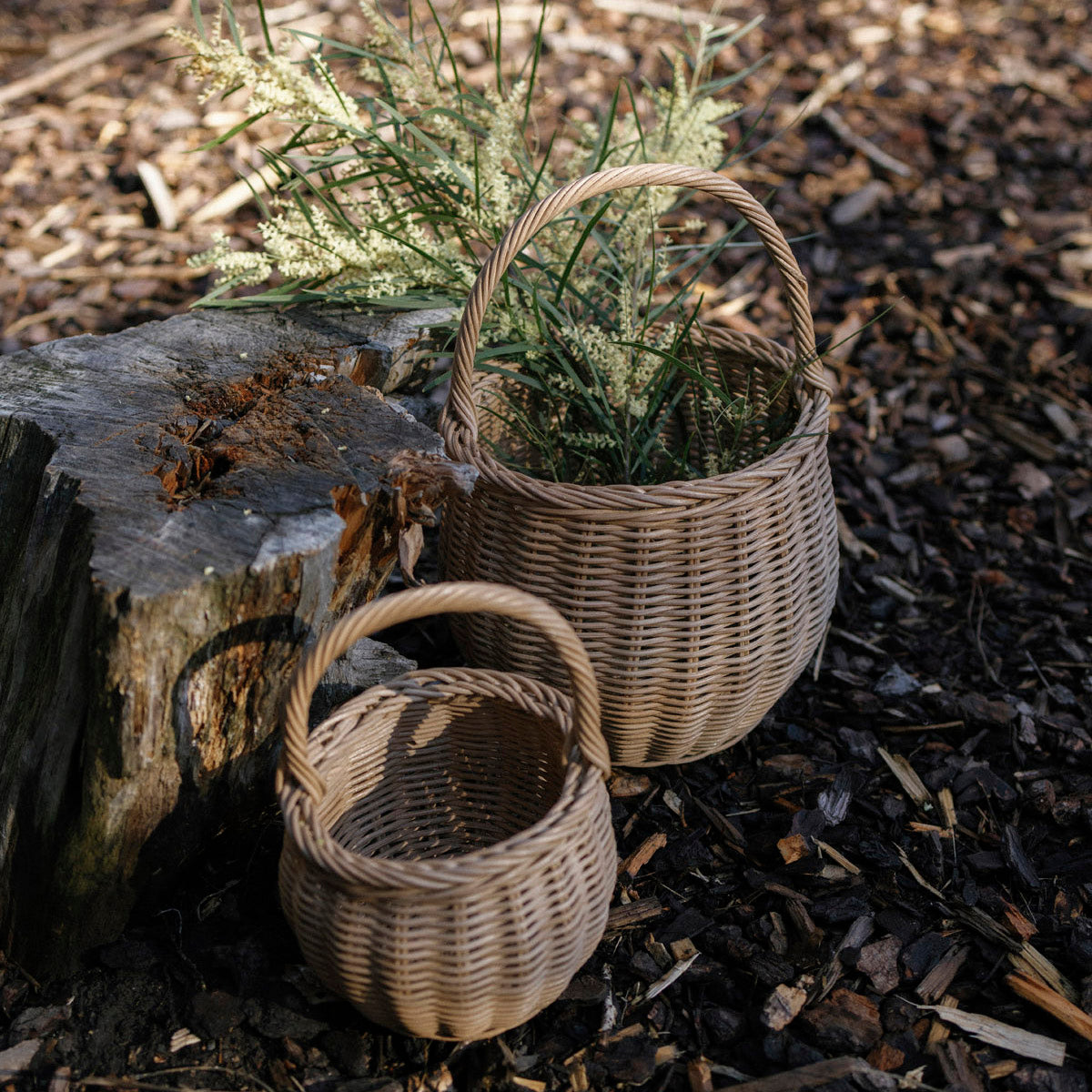 Two small woven wicker baskets with handles, one large and one small, placed outdoors on mulch beside a weathered log with dried grass.