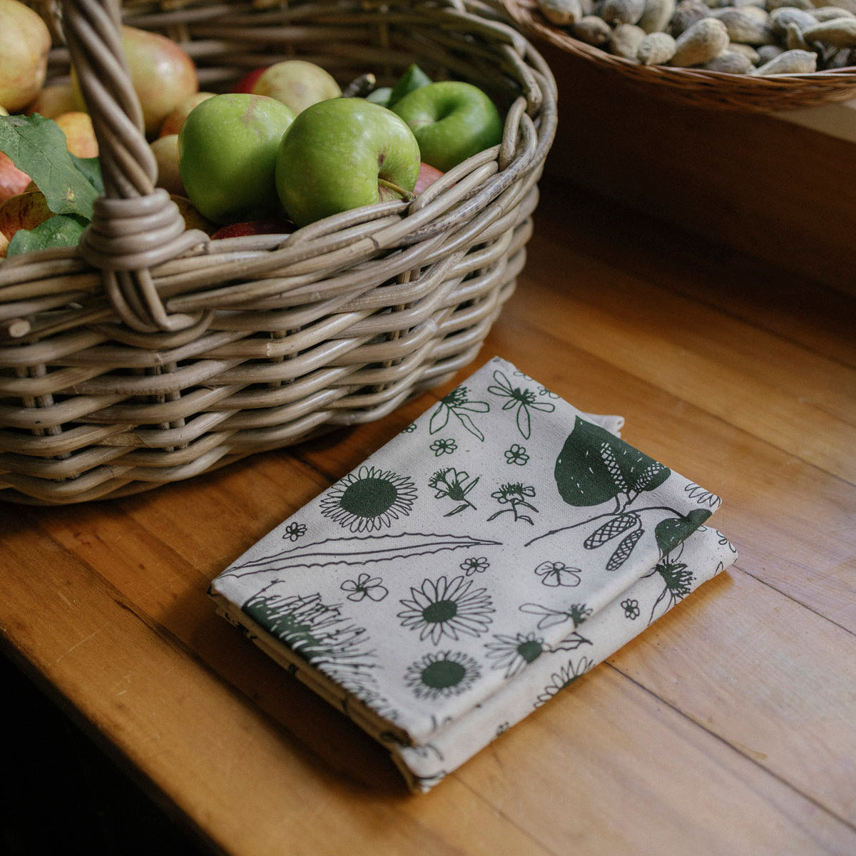 Wicker basket with apples on a wooden surface next to folded fabric with nature-themed design