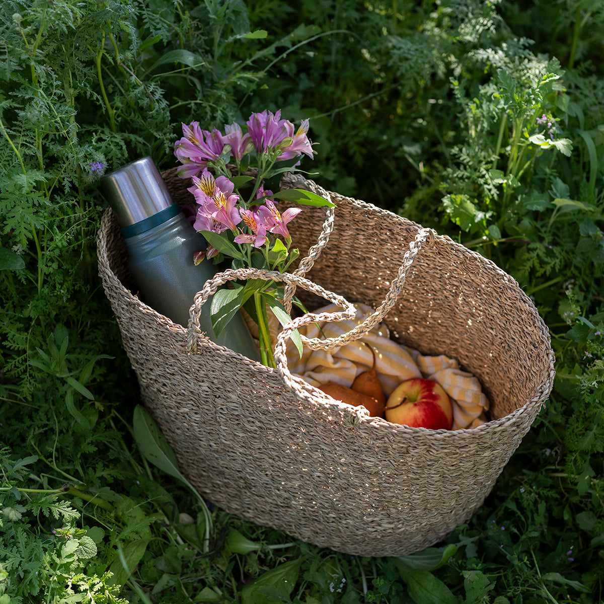 Picnic basket with apples, flowers, and a thermos in a grassy setting. 