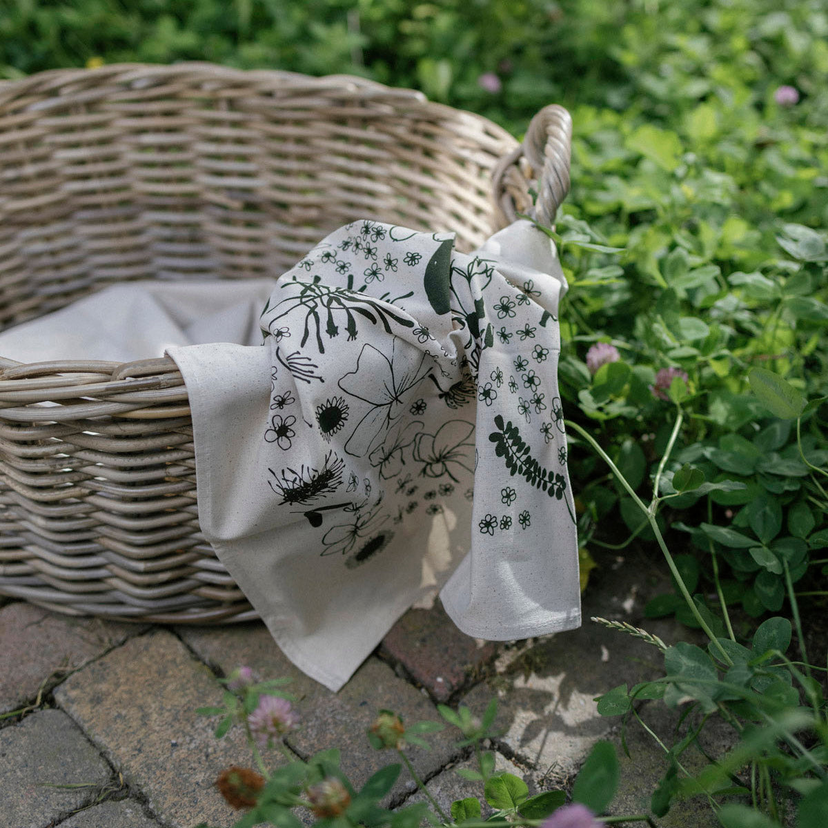 Wicker basket with a floral-patterned white towel on a stone path with greenery.