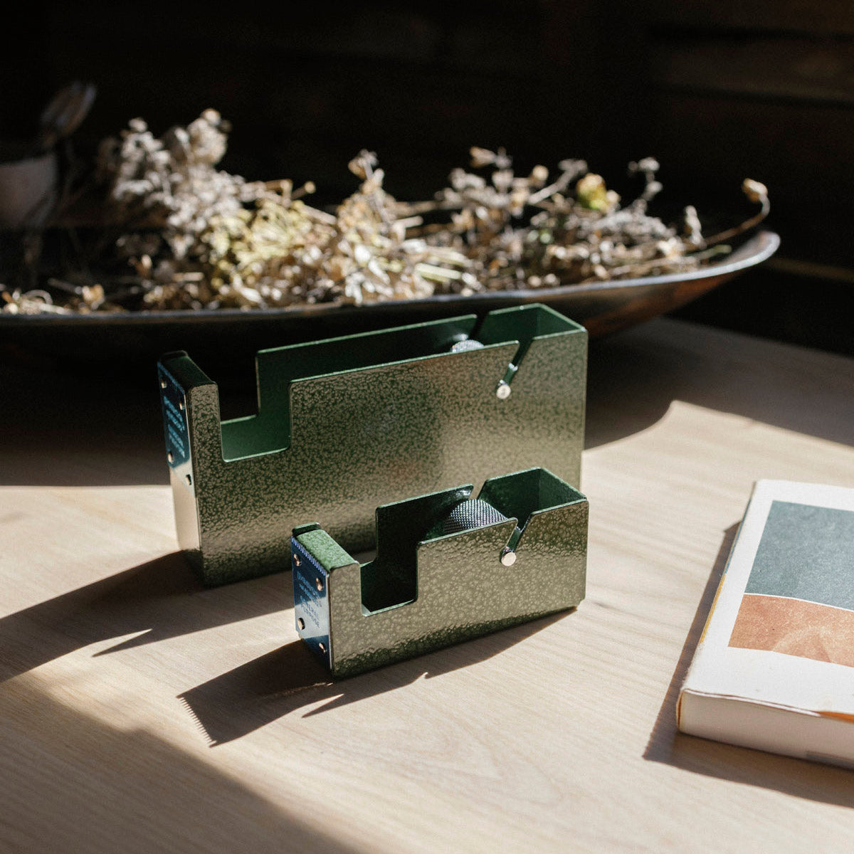 Two green metal tape dispensers on a wooden surface with a bowl of dried plants in the background.