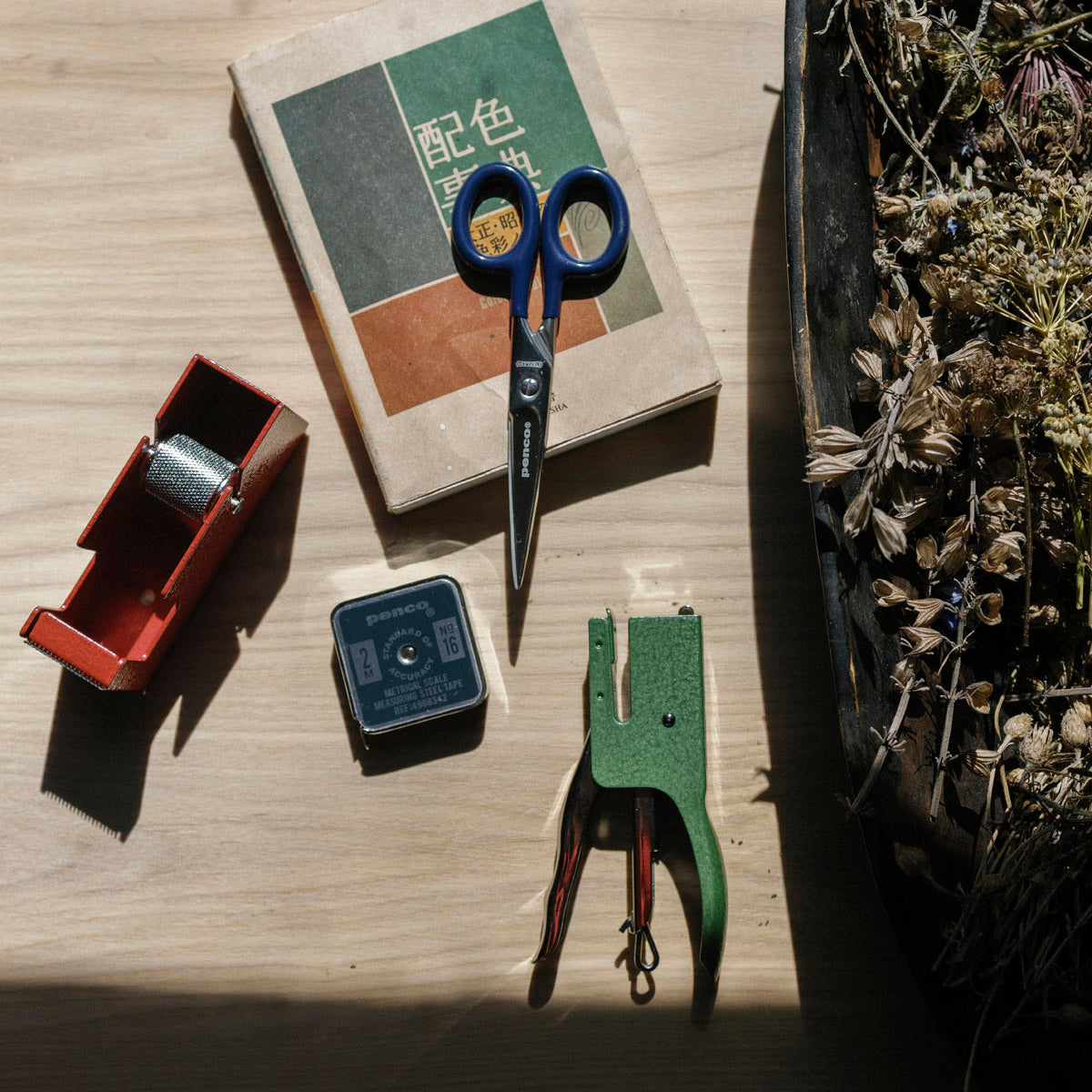 Scissors, a small metal measuring tape, a tape dispenser and a stapler on a wooden surface with a book and plant in the background.