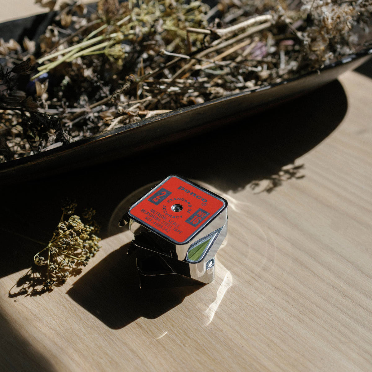 Three small Penco Tape Measures with labels on a wooden surface, surrounded by dried plants.
