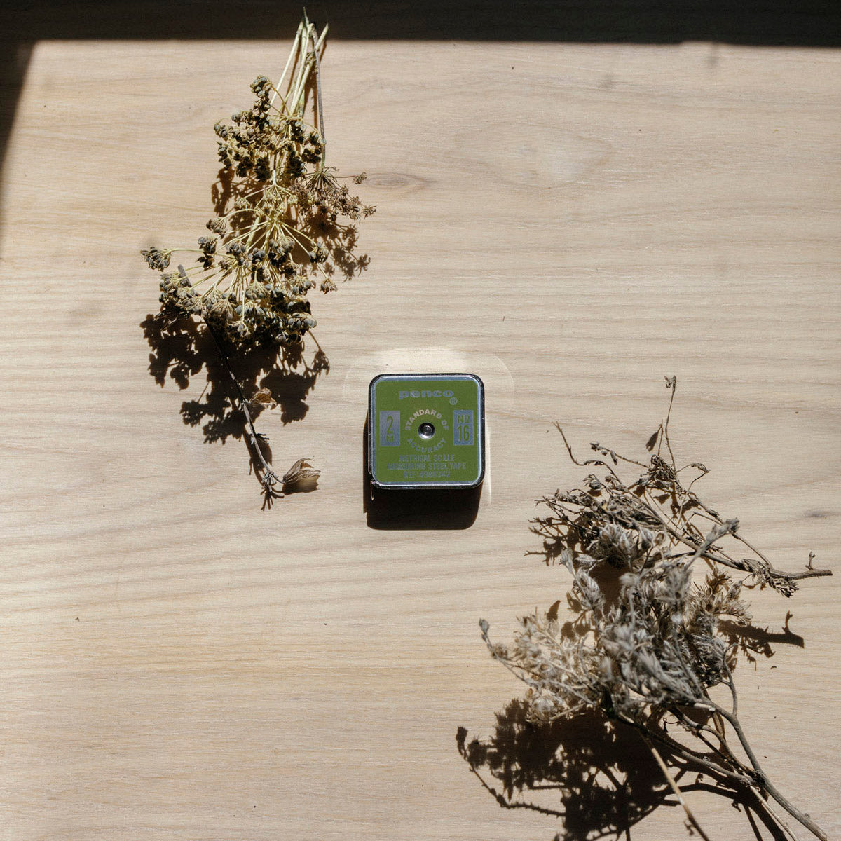 Small square metal tape measure with a green label on a wooden surface with dried plants.