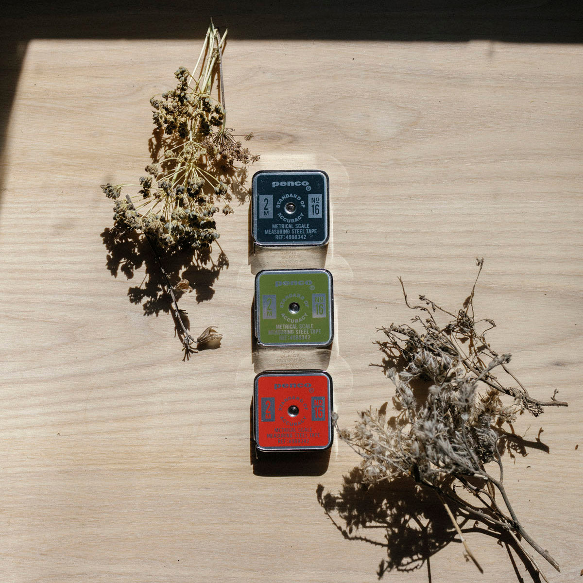 Three small Penco Tape Measures with labels on a wooden surface, surrounded by dried plants.