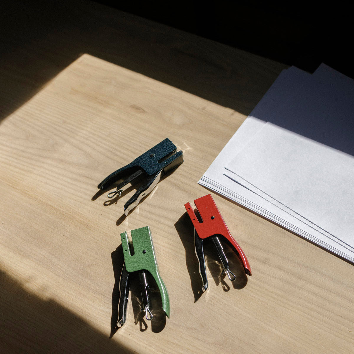 Three colorful industrial steel staplers on a wooden surface with a stack of white paper.
