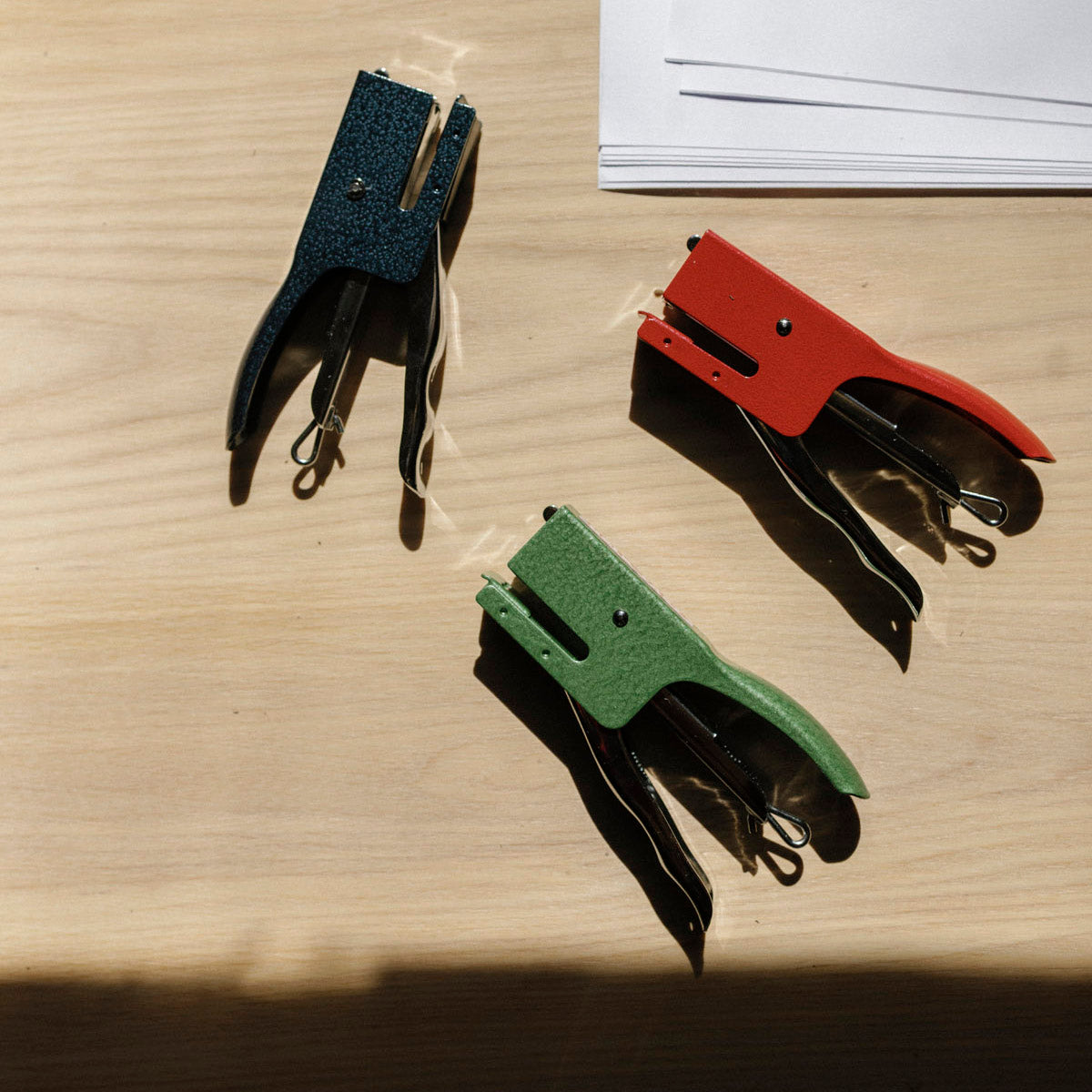 Three colorful staplers on a wooden surface with a stack of paper in the background.