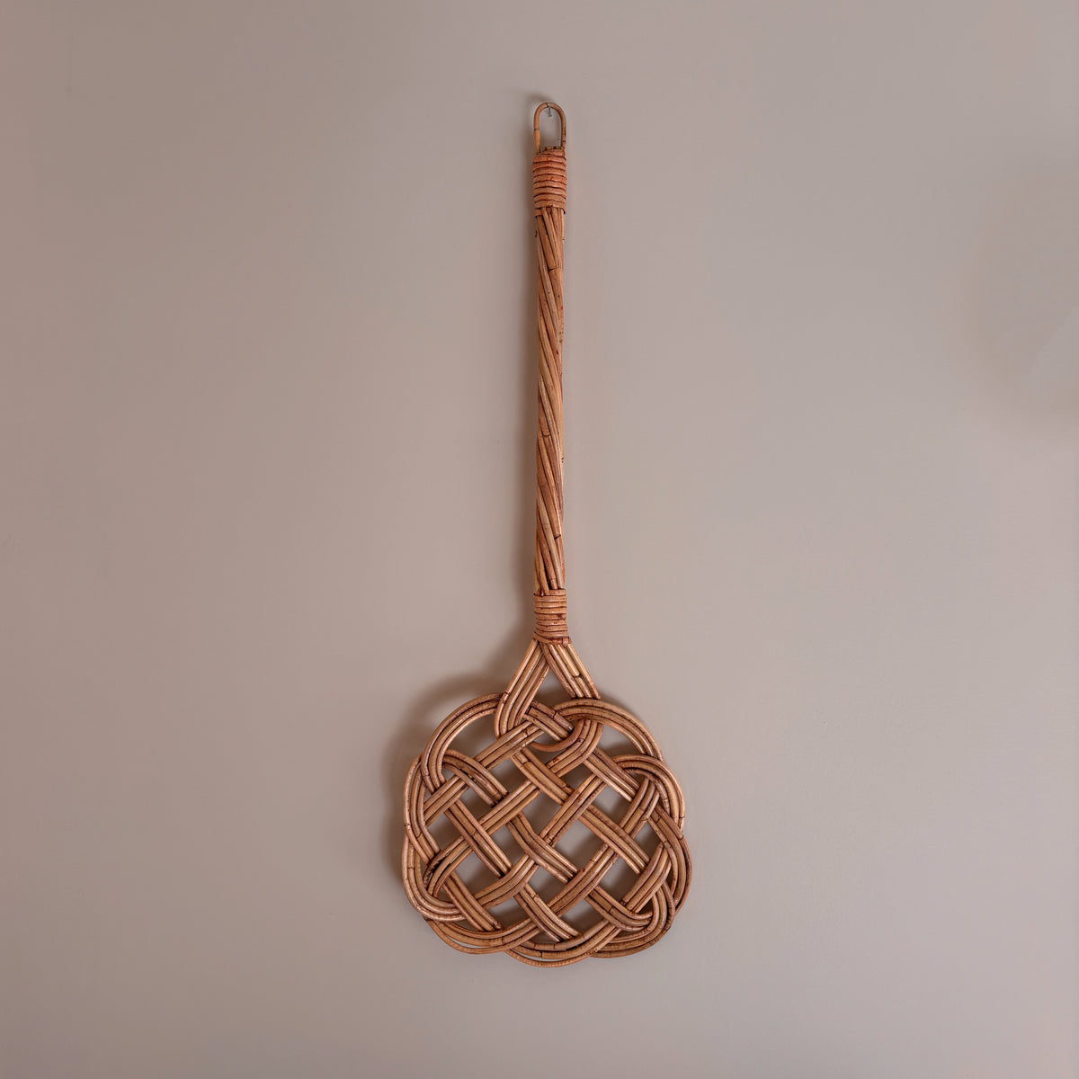 A woven rattan wall hook with a long handle and a decorative interlaced oval head, hanging against a plain light beige wall.