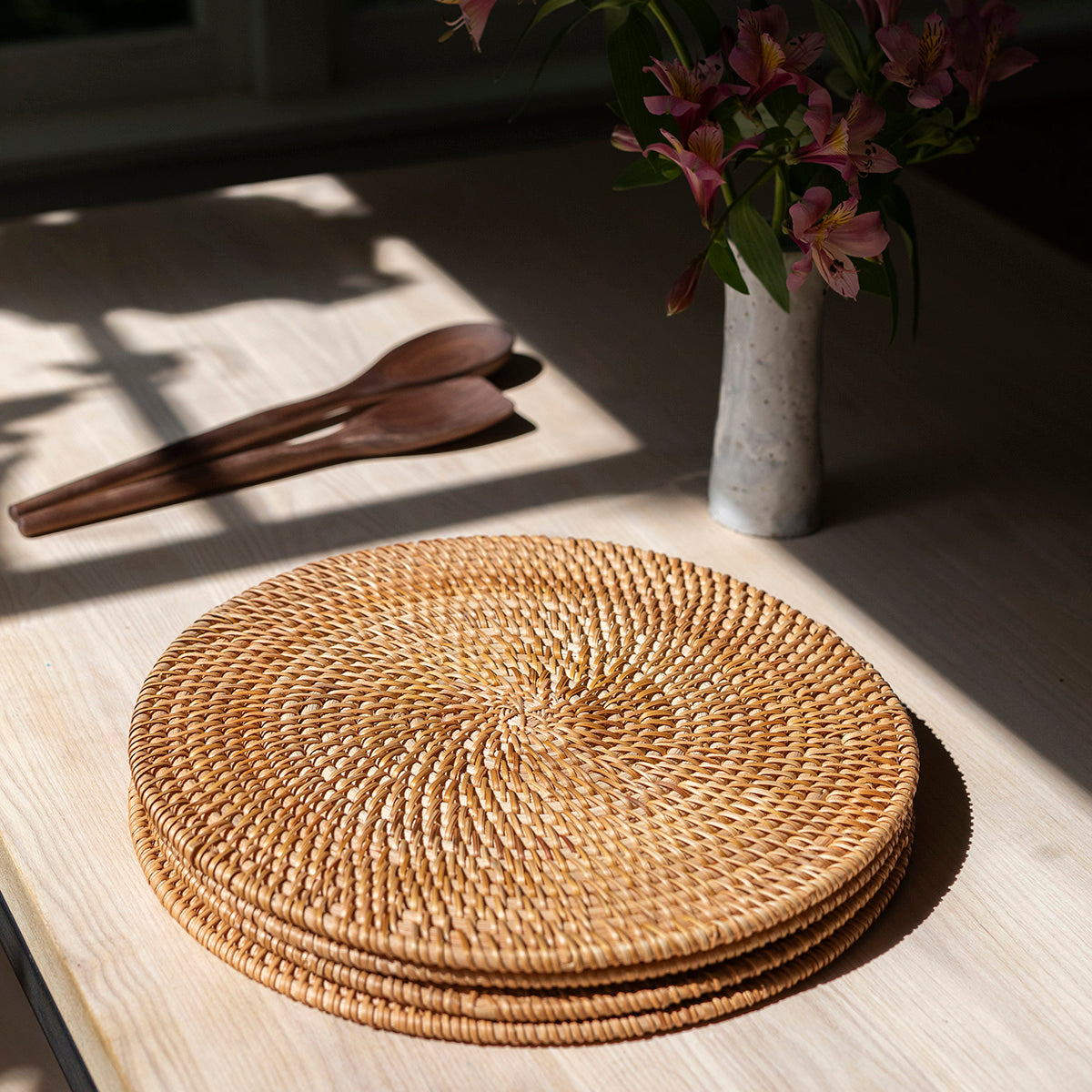 Stack of woven placemats on a wooden surface with a vase of flowers in the background.