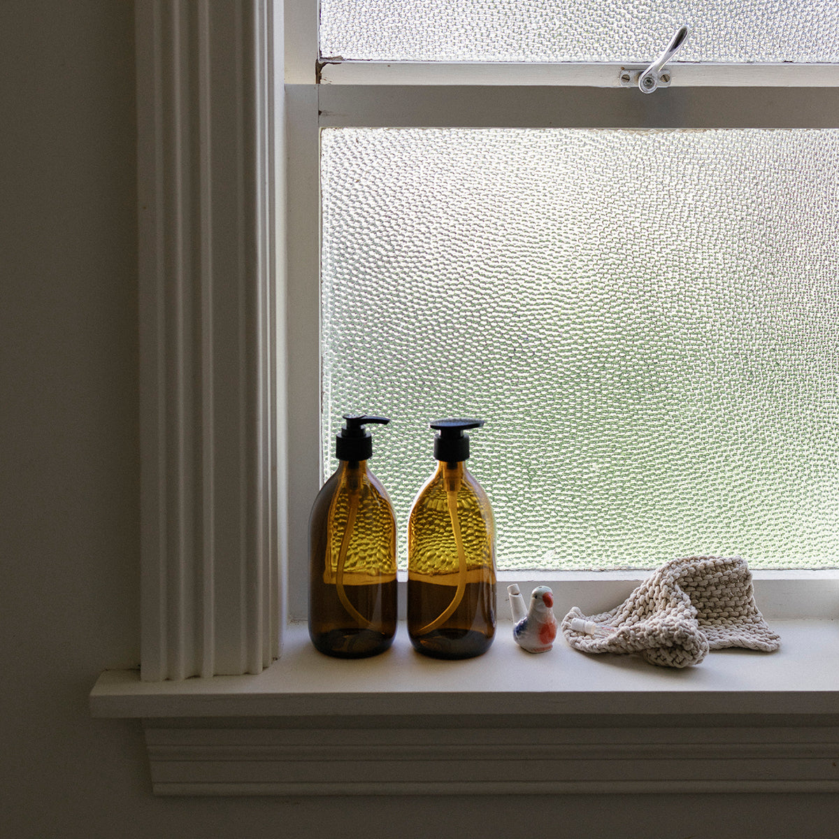 Two glass bottles with black pumps on a windowsill with a textured glass window.
