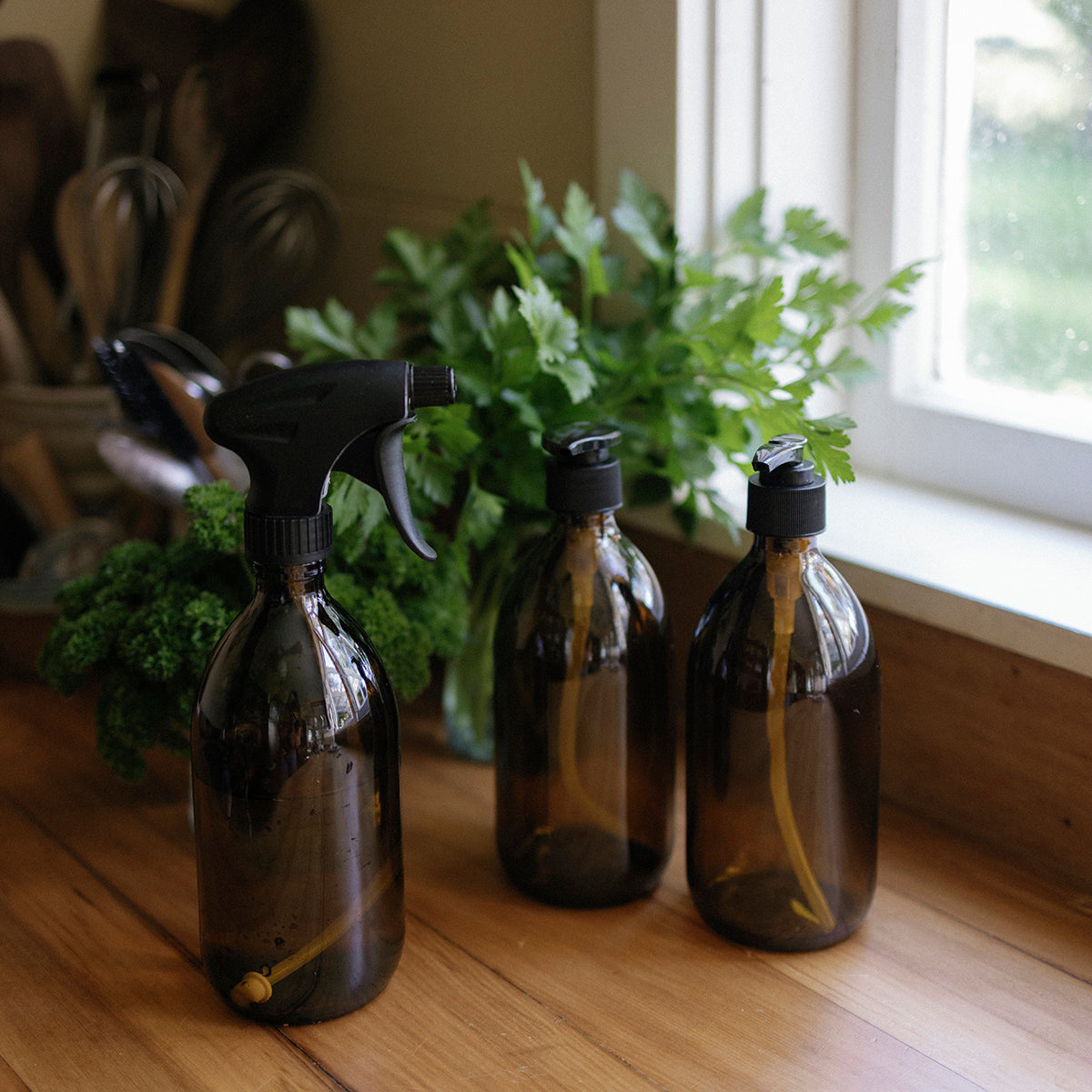 Three brown glass bottles with black sprayers on a wooden surface near a window with plants.