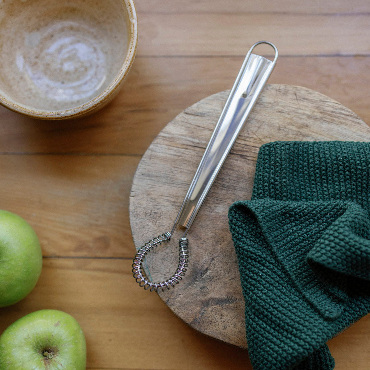 Metallic whisk on a wooden board with green apples and a bowl in the background