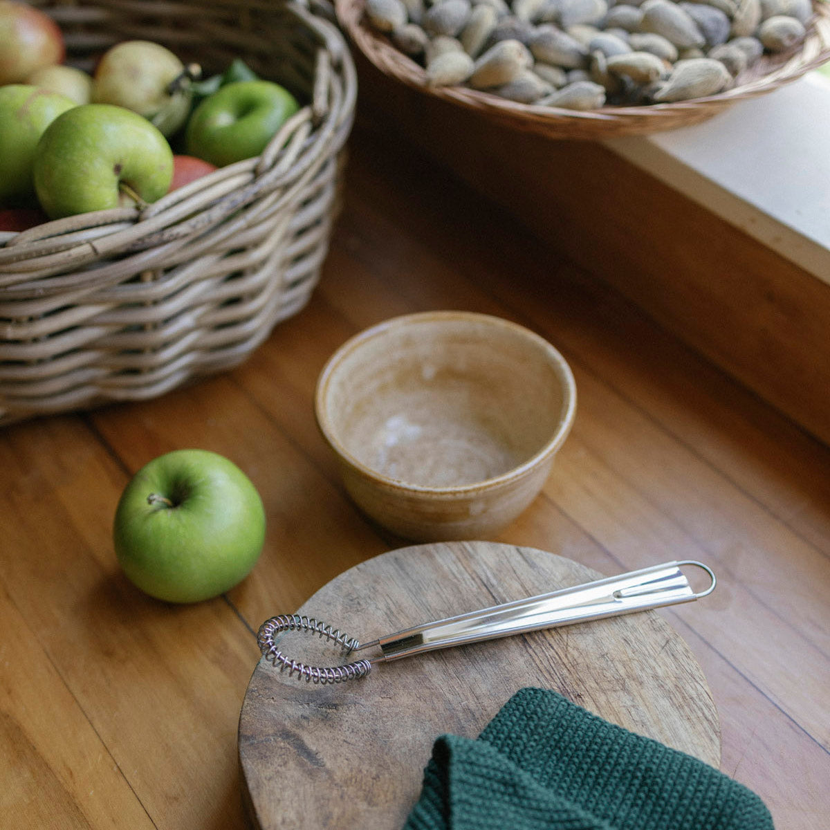 Green apple on a wooden surface with a ceramic bowl, metal whisk, and basket of apples in the background.