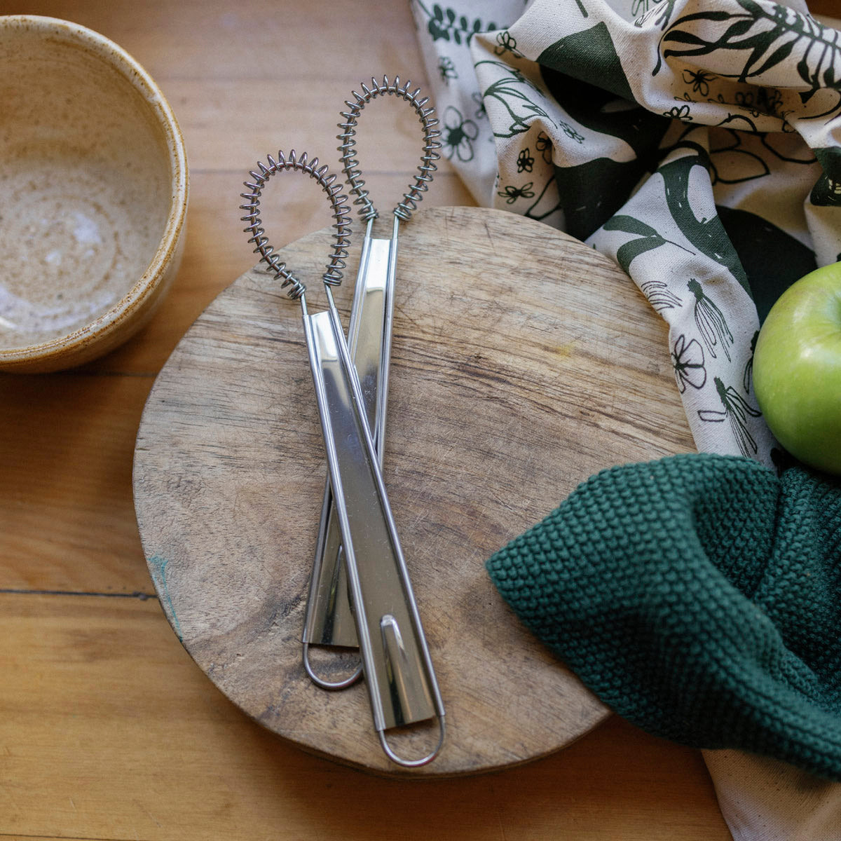 Spiral whisk kitchen tool on a wooden board with a green apple and patterned cloth in the background.